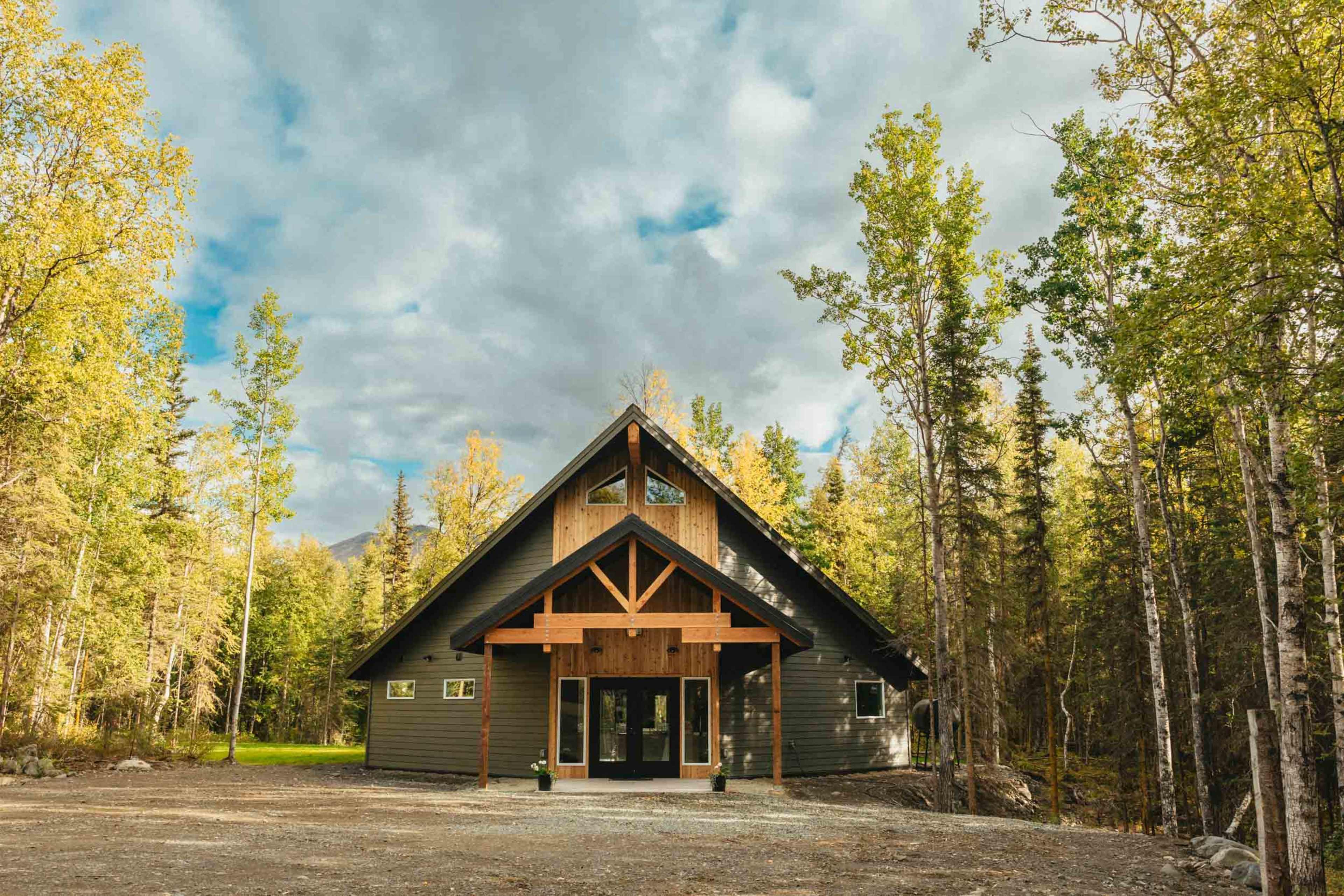 A modern cabin with a peaked roof and wooden accents is surrounded by tall trees and a cloudy sky.
