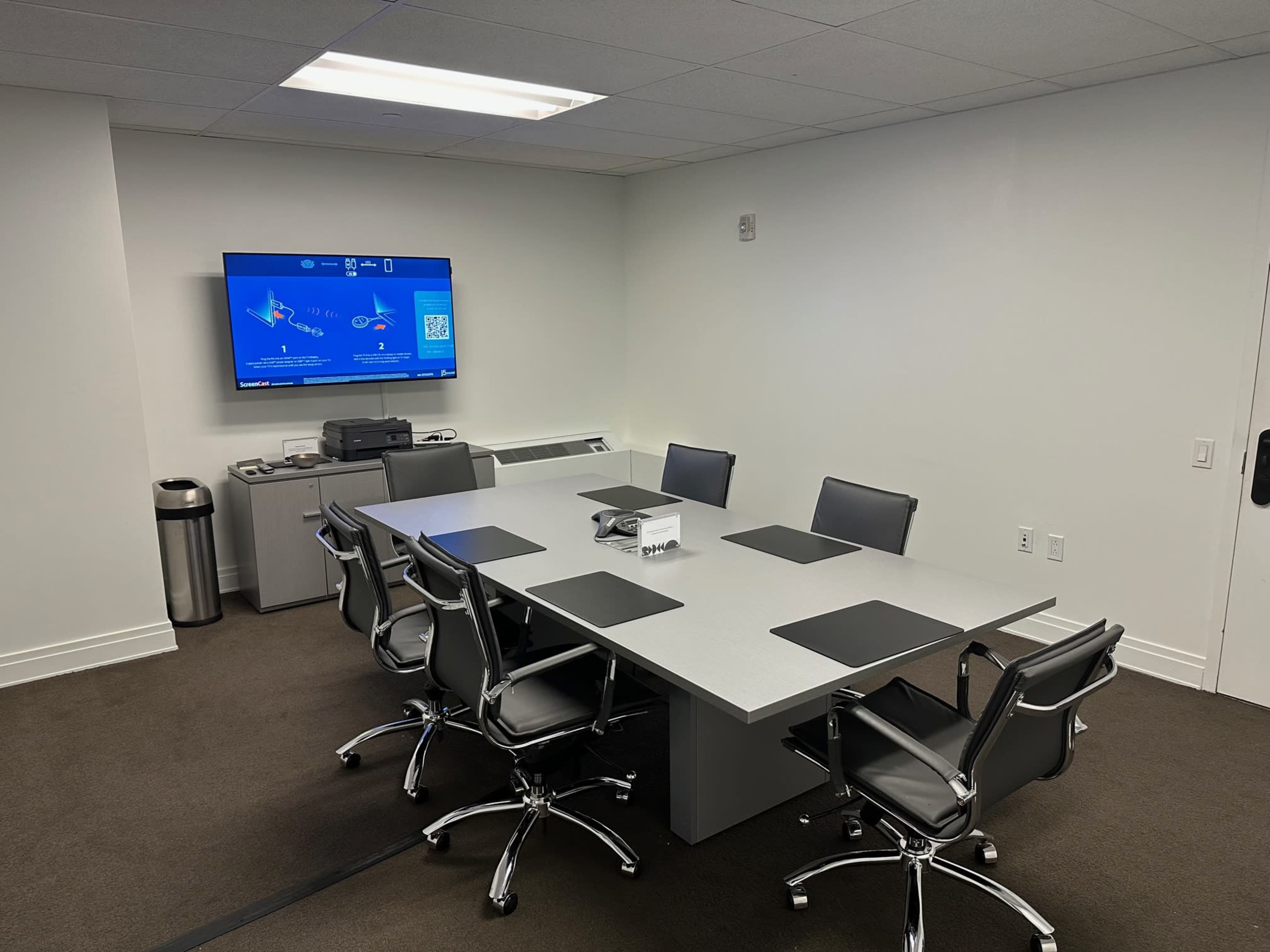 A conference room is equipped with a large table, six chairs, and a wall-mounted screen displaying a presentation.