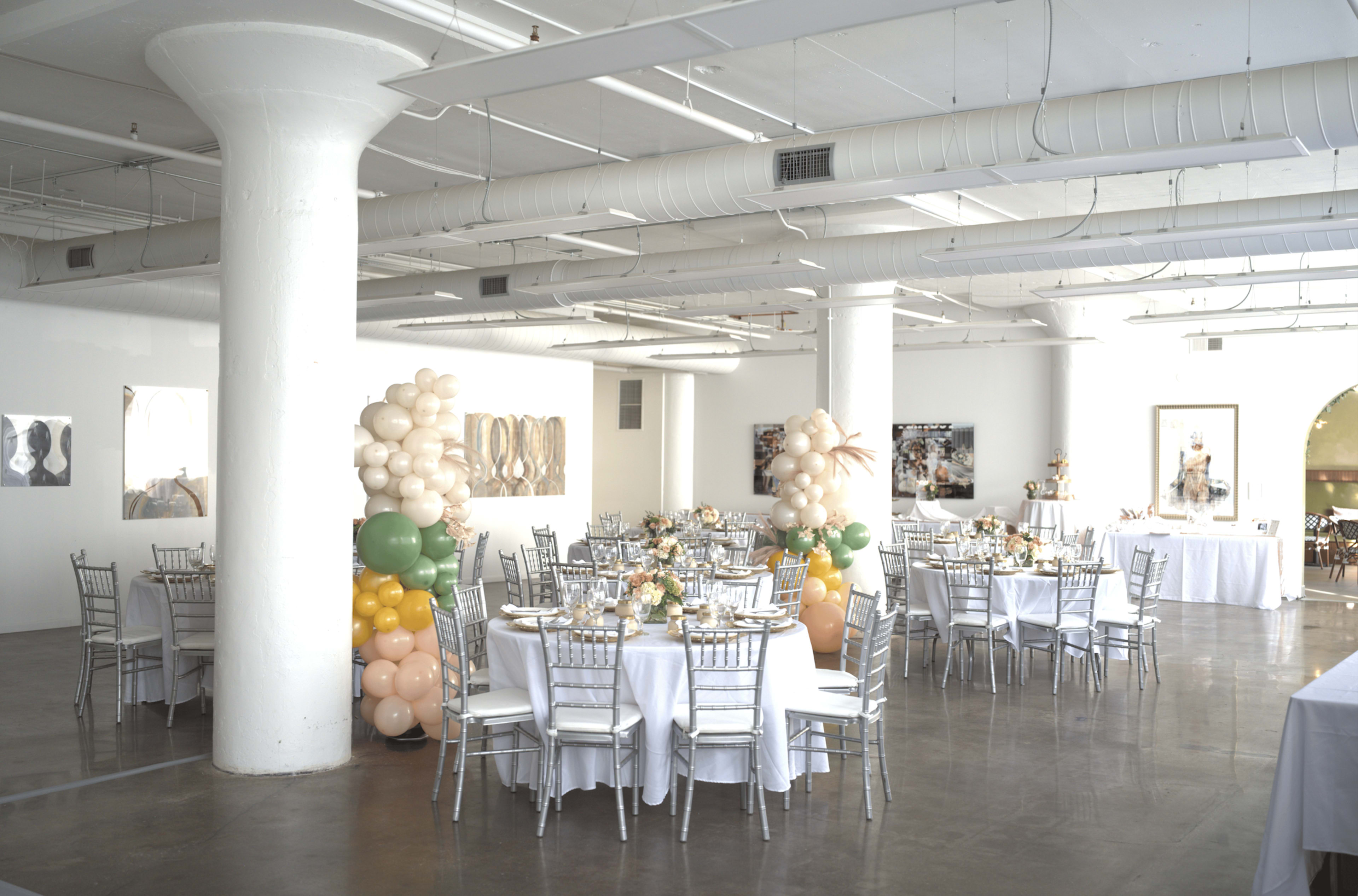 The image shows a spacious banquet hall set up for an event, featuring tables with white linens and centerpieces, alongside decorative balloon arrangements in the corners.
