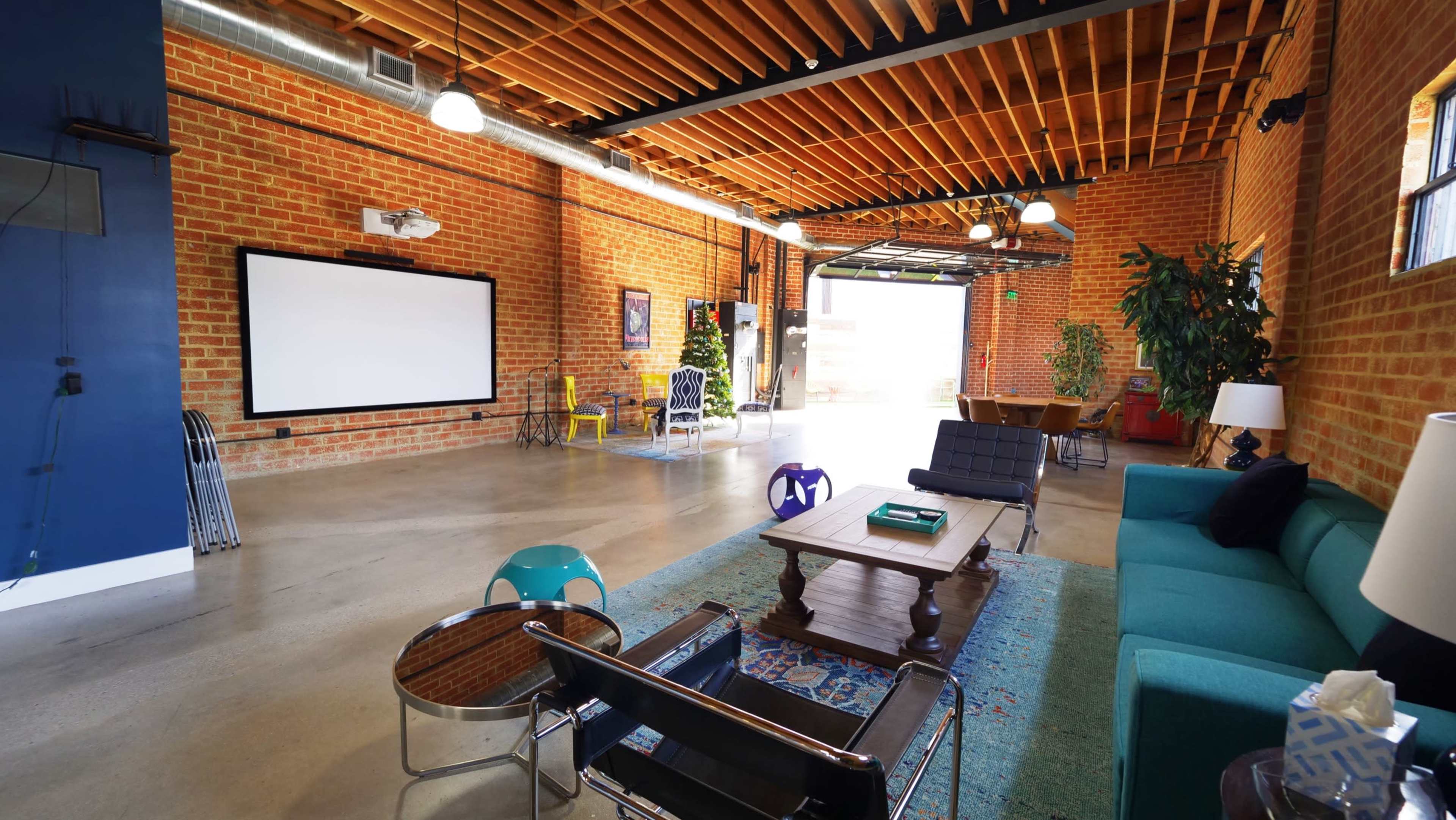 The image shows an industrial-style interior with exposed brick walls, a large white screen, and various seating arrangements.