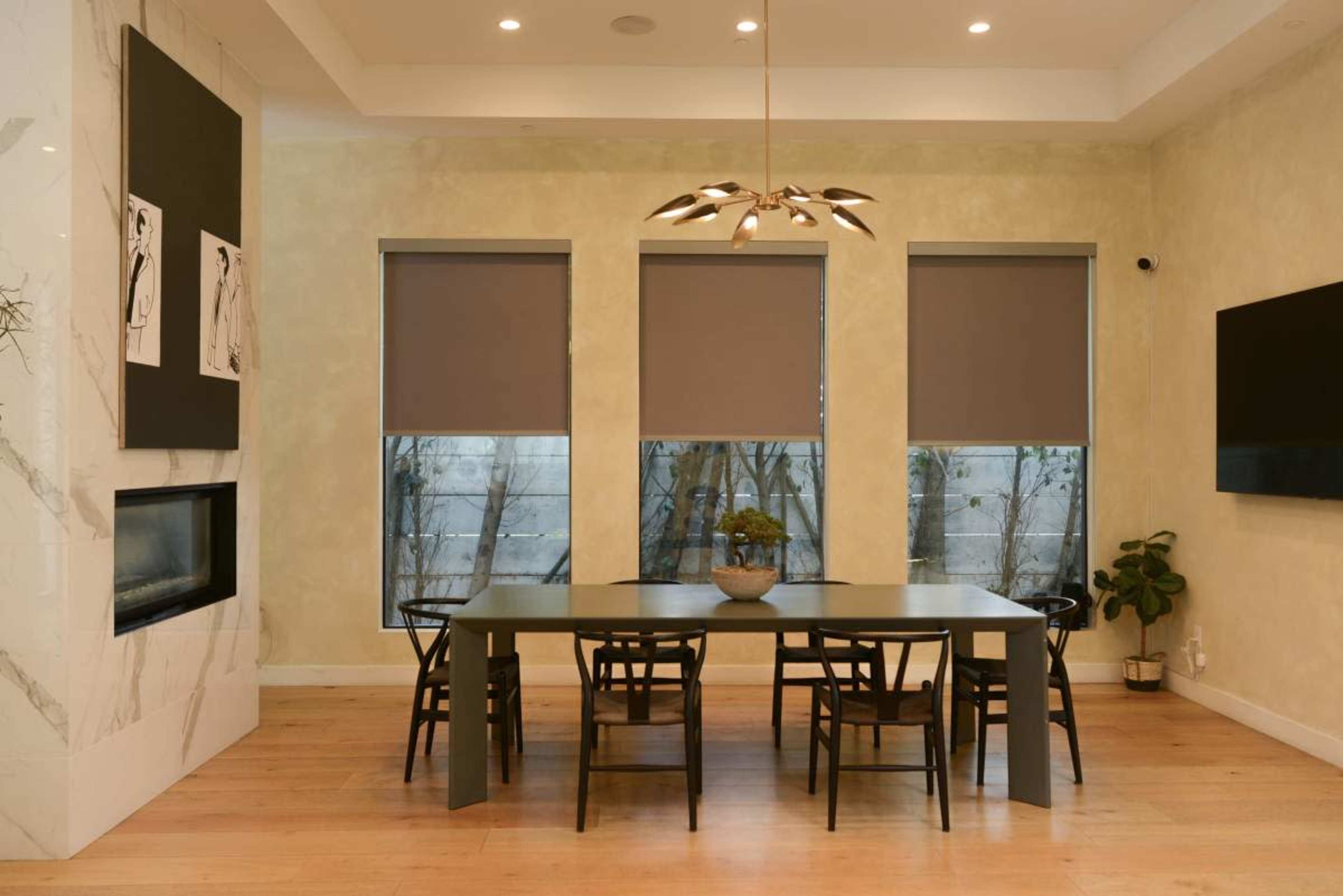 A modern dining area features a long table surrounded by black chairs, with three large windows covered by gray shades and a mounted television on the wall.