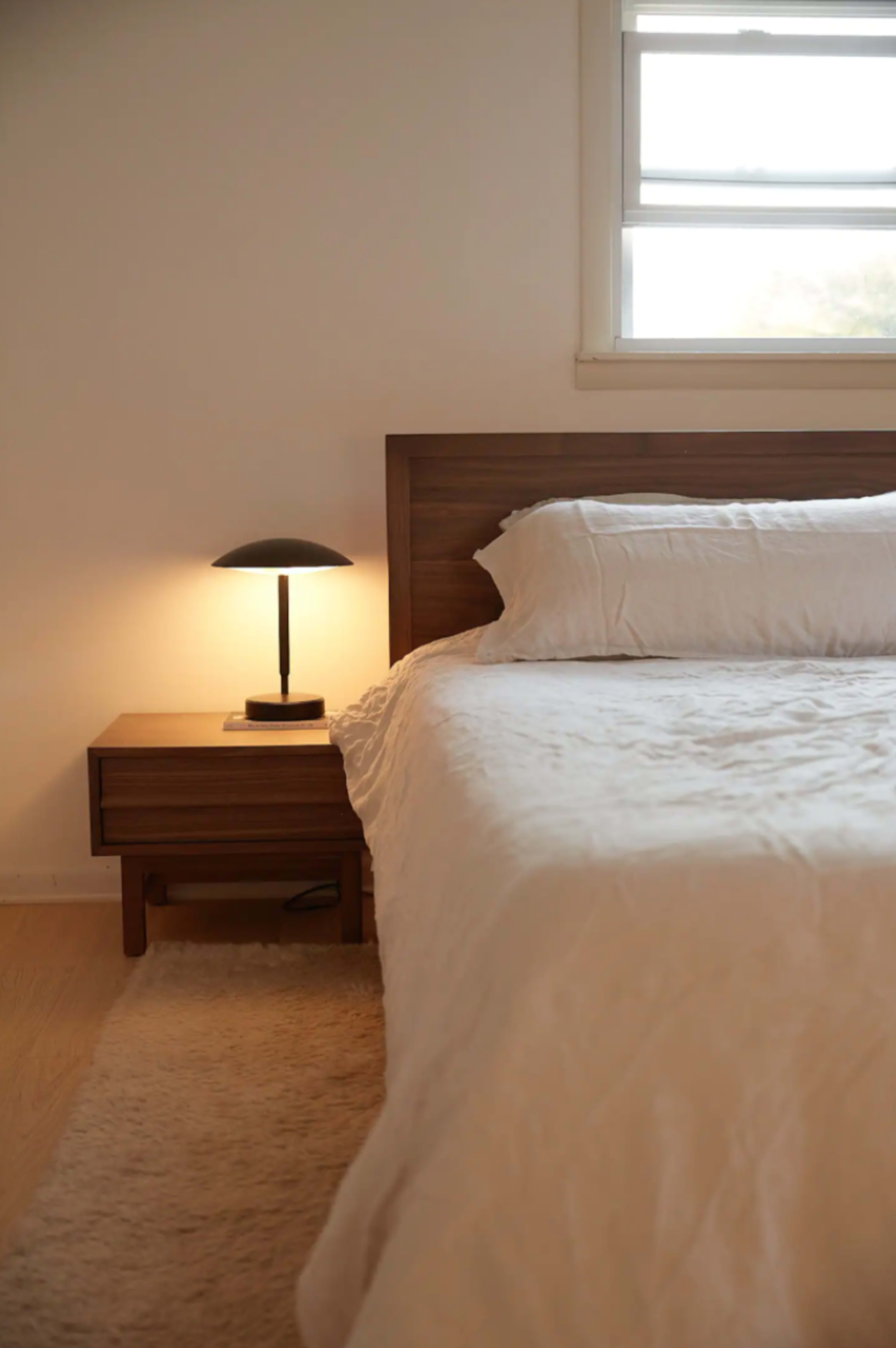 A neatly made bed with white bedding is positioned next to a wooden nightstand and a lamp emitting a warm light.