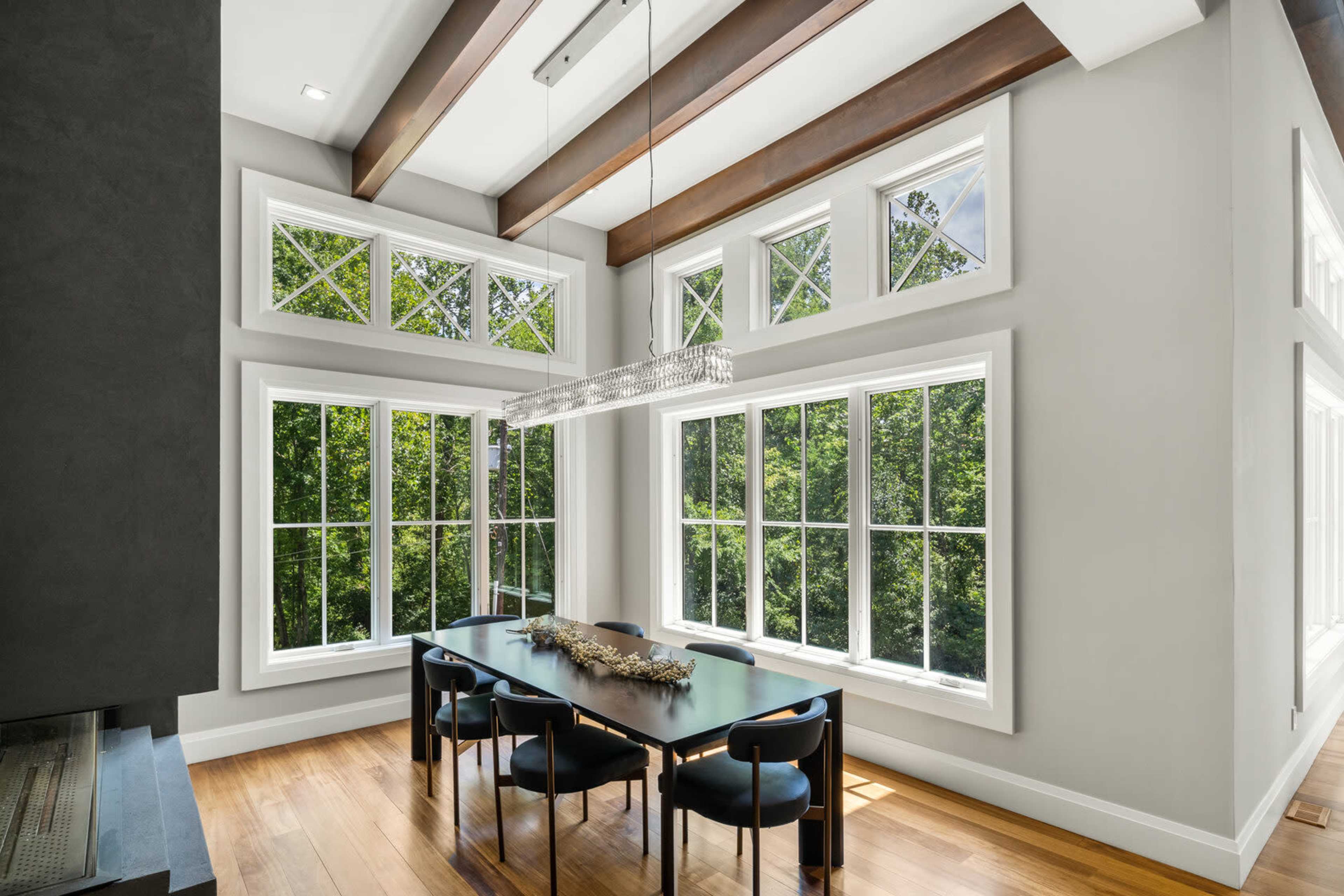 A dining area features a dark wood table surrounded by black chairs, illuminated by a modern light fixture, with large windows that offer a view of greenery outside.
