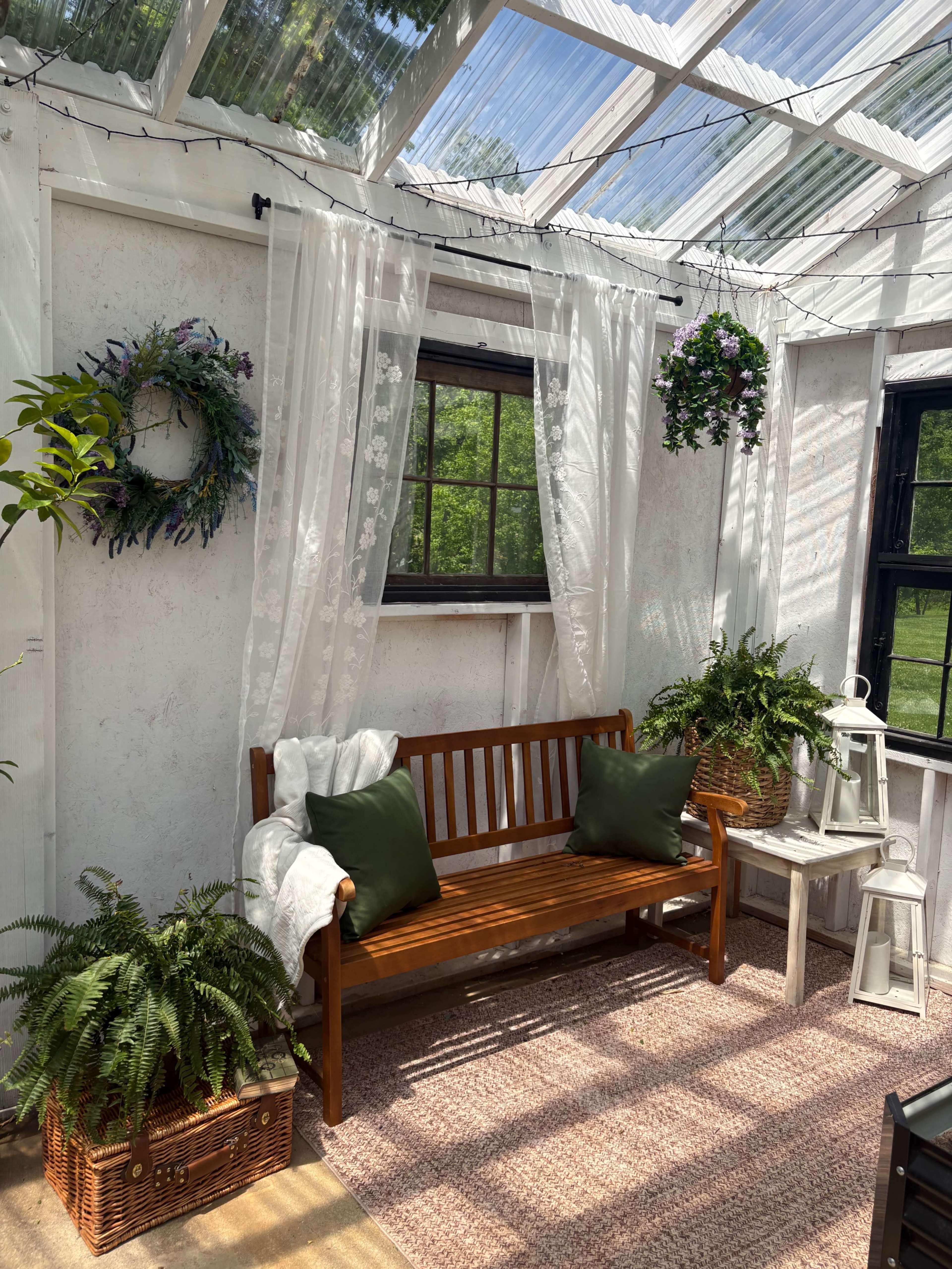 The image shows a sunlit indoor space with a wooden bench, potted plants, sheer curtains, and decorative elements like a wreath and hanging flowers.