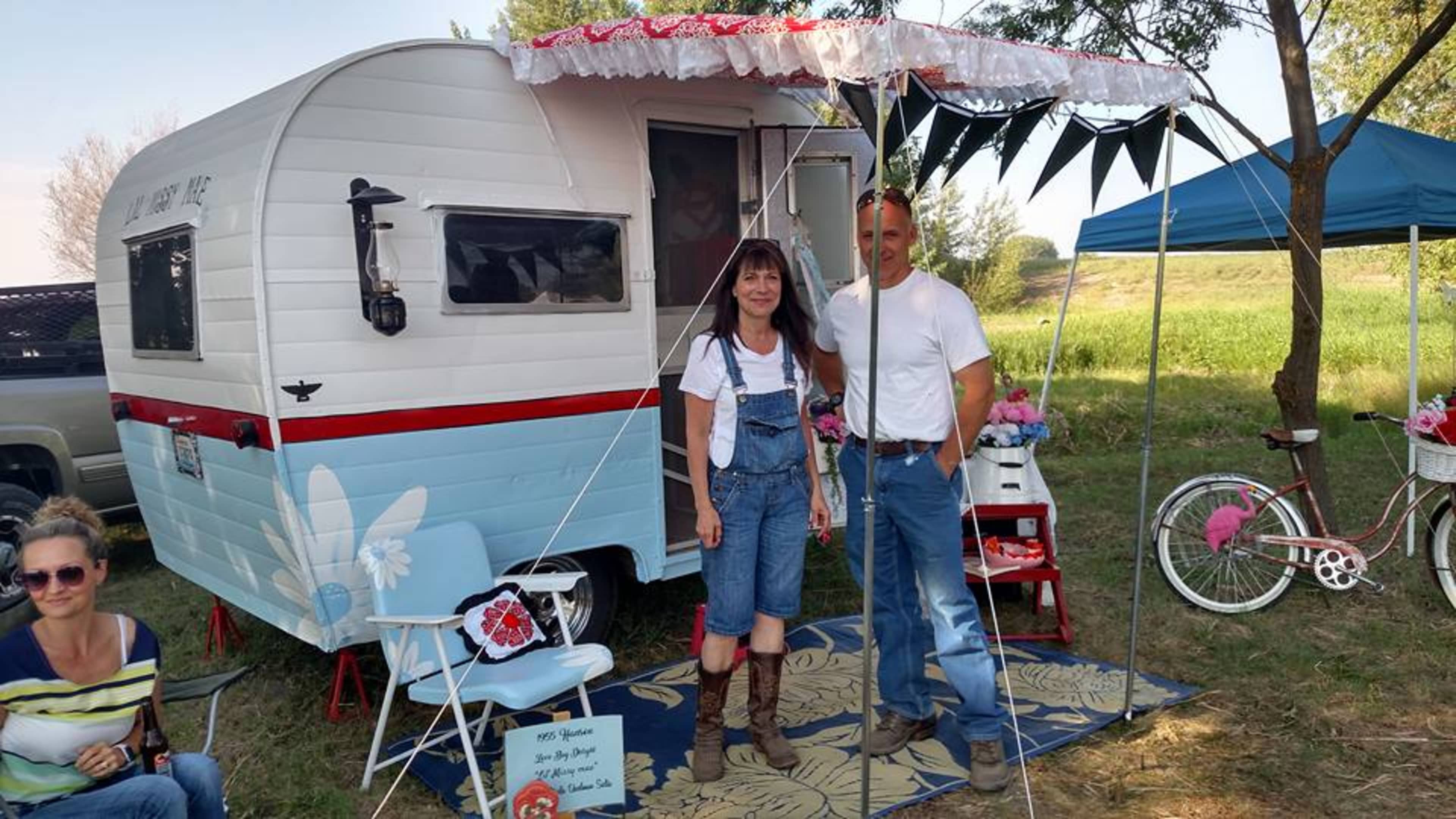 A man and a woman stand in front of a parked vintage camper adorned with decorations, while a bicycle and chairs are nearby in a grassy area.