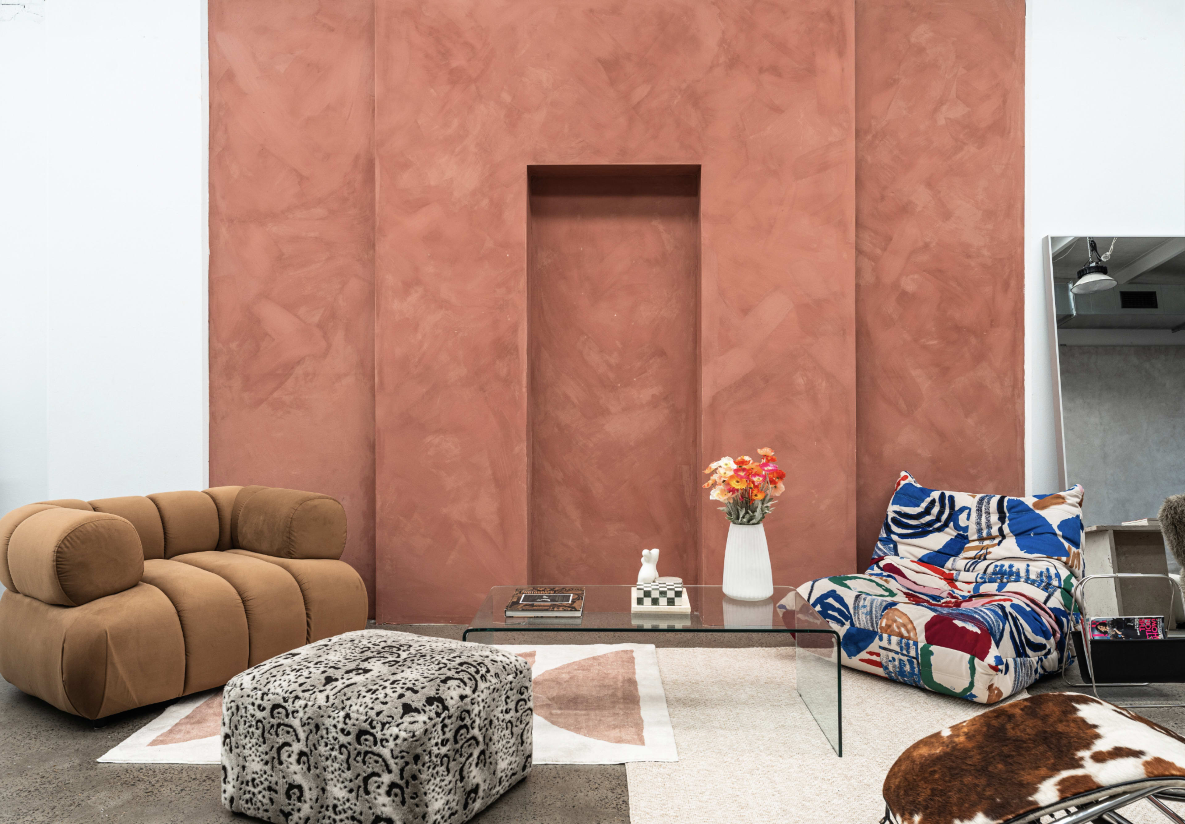 The image shows a modern living room featuring two unique seating arrangements, a geometric coffee table, and a flower arrangement against a rust-colored wall.