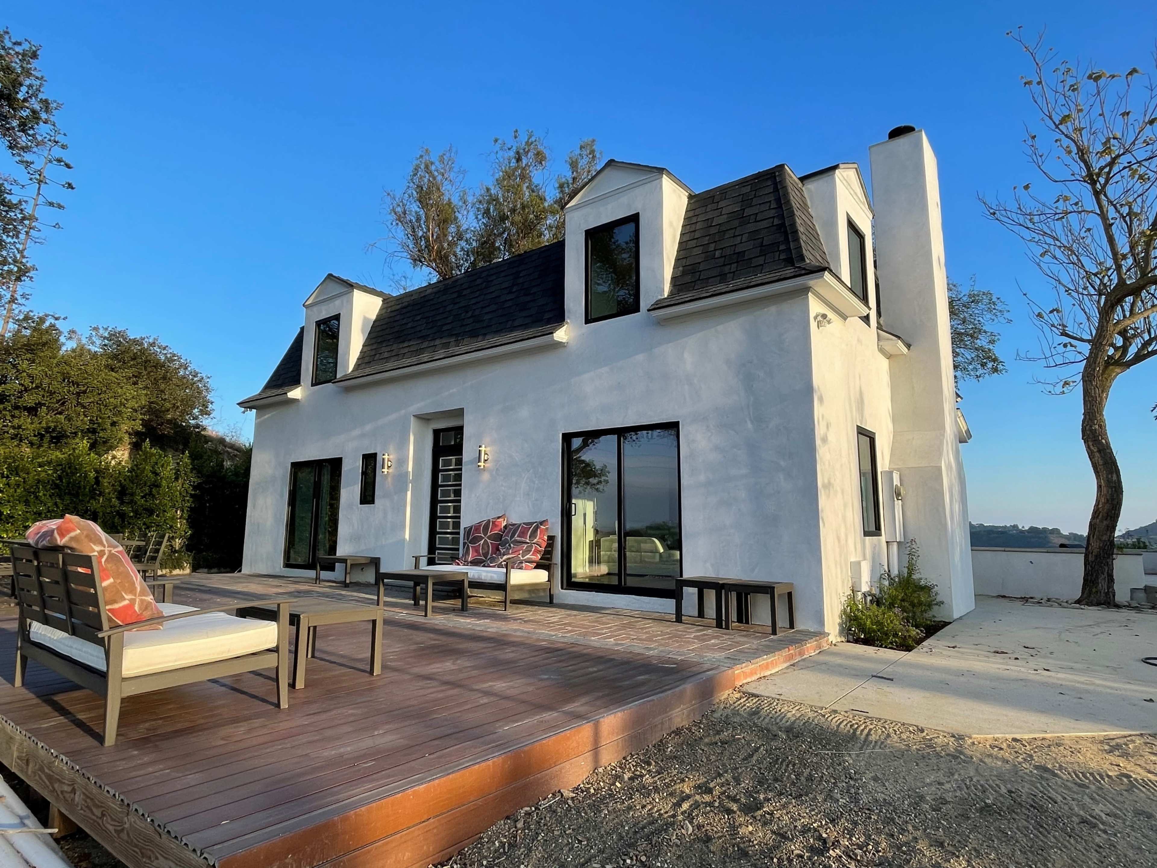 A modern two-story house with a sloped roof and large windows is situated next to a wooden deck and a leafless tree.