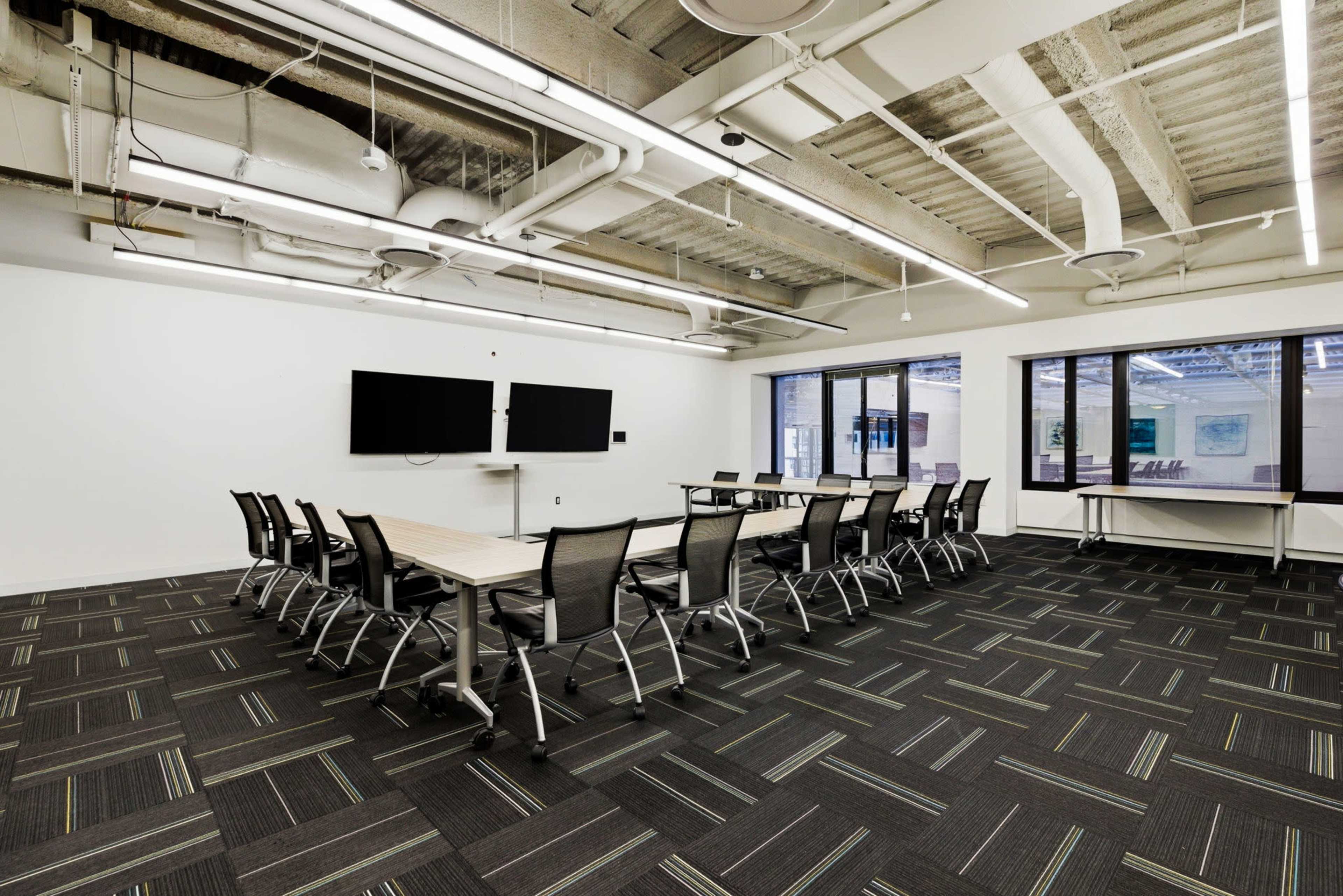 The image shows a modern conference room with a long table set for meetings, surrounded by black mesh chairs, and equipped with two wall-mounted screens.