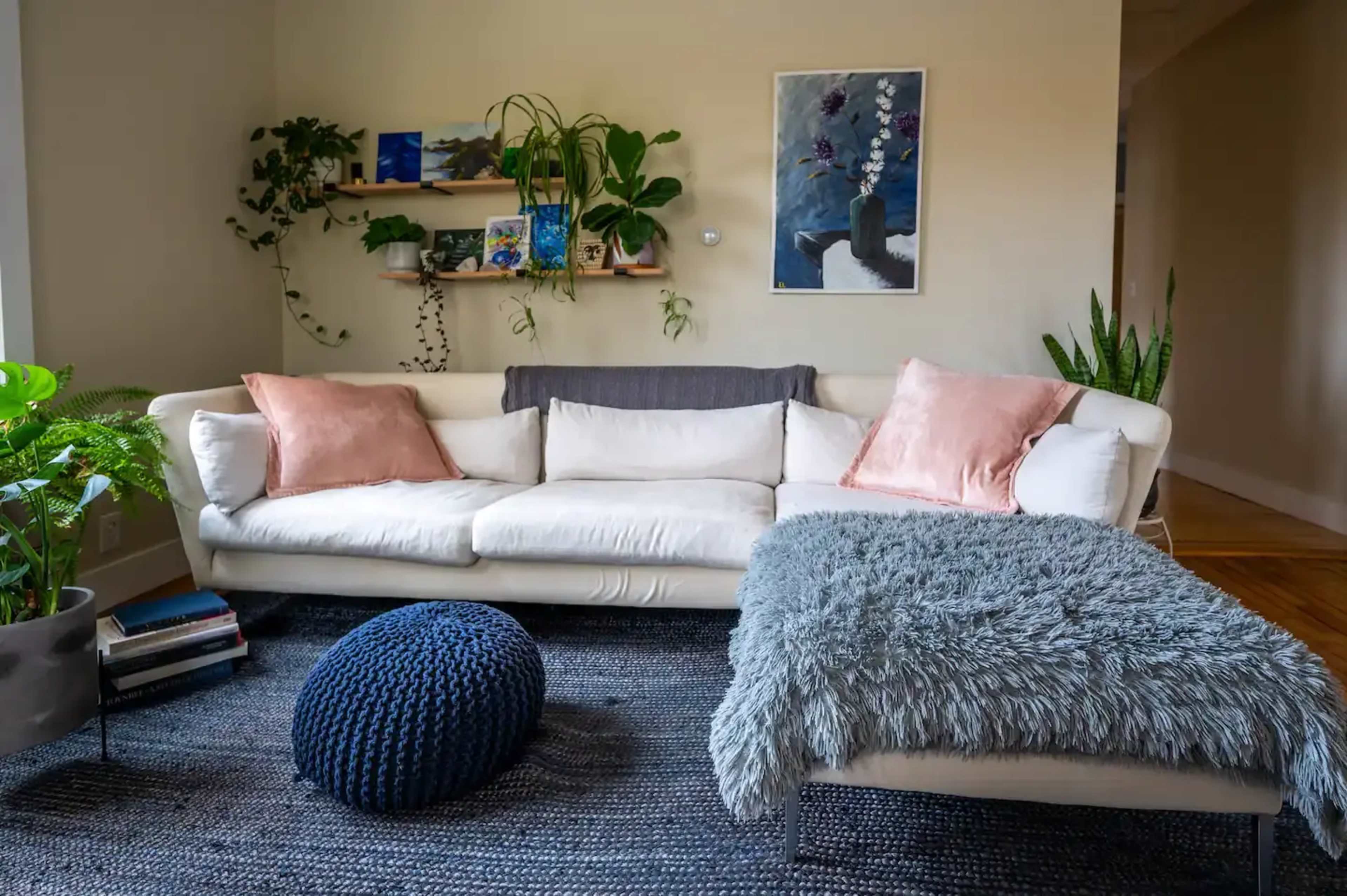 A bright living room features a white sectional sofa with pink pillows, a textured gray throw, and a blue pouf, surrounded by plants and artwork on the walls.