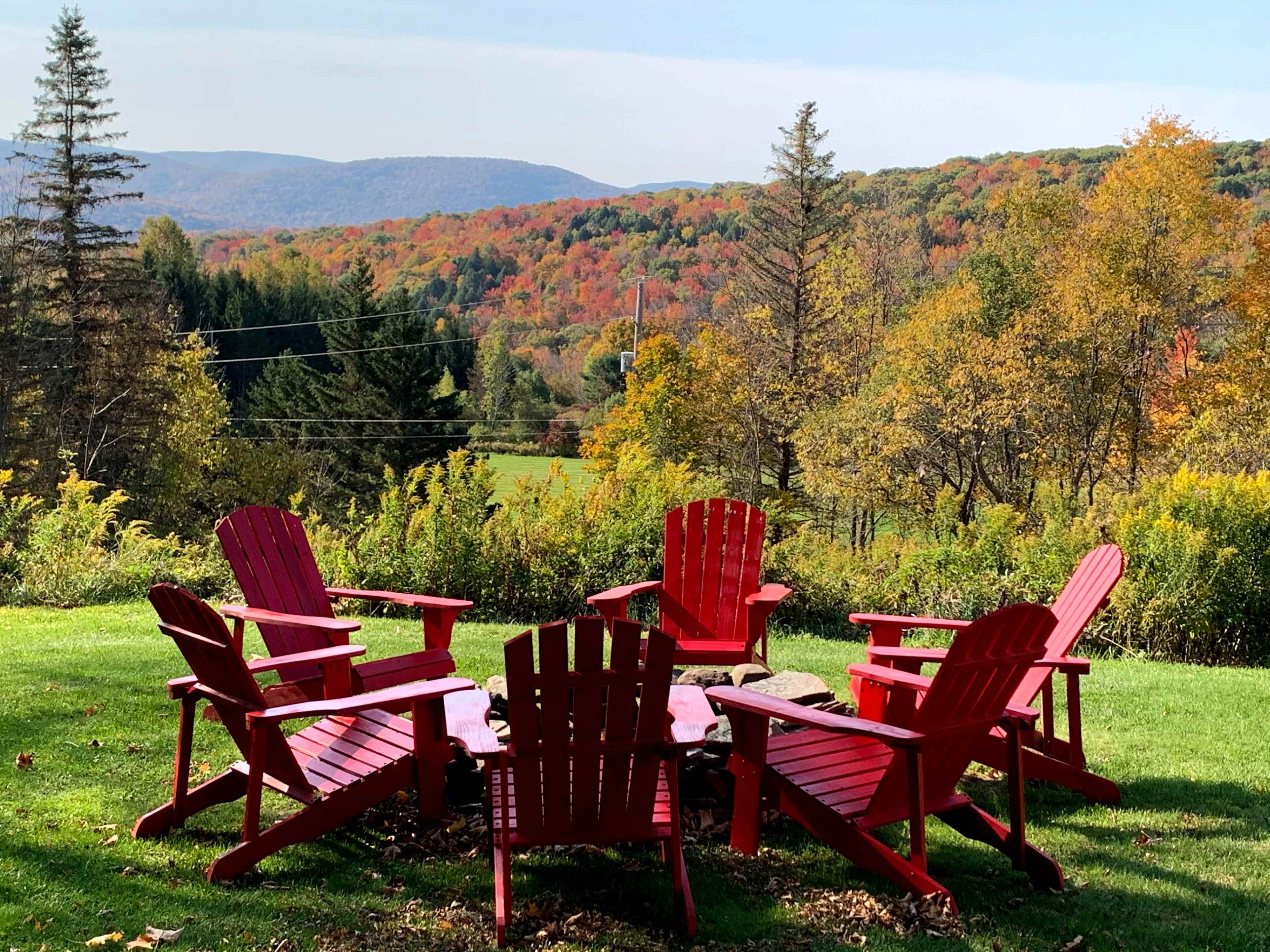 A circle of six red Adirondack chairs surrounds a wooden table in an open green space, backed by a colorful autumn landscape of rolling hills and trees.