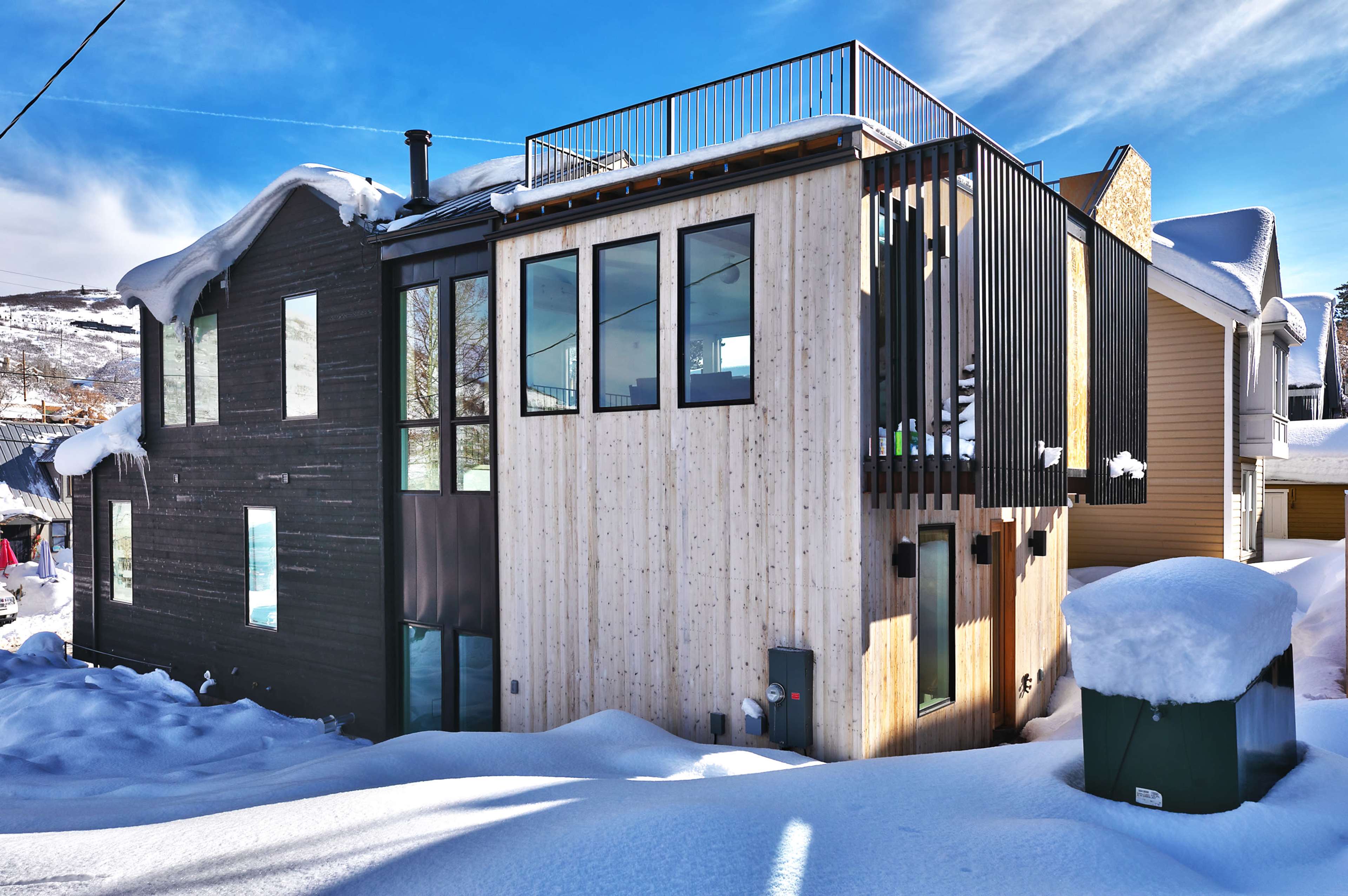 A modern house with a mix of wooden and dark exterior finishes is partially buried in snow, featuring large windows and a rooftop terrace.