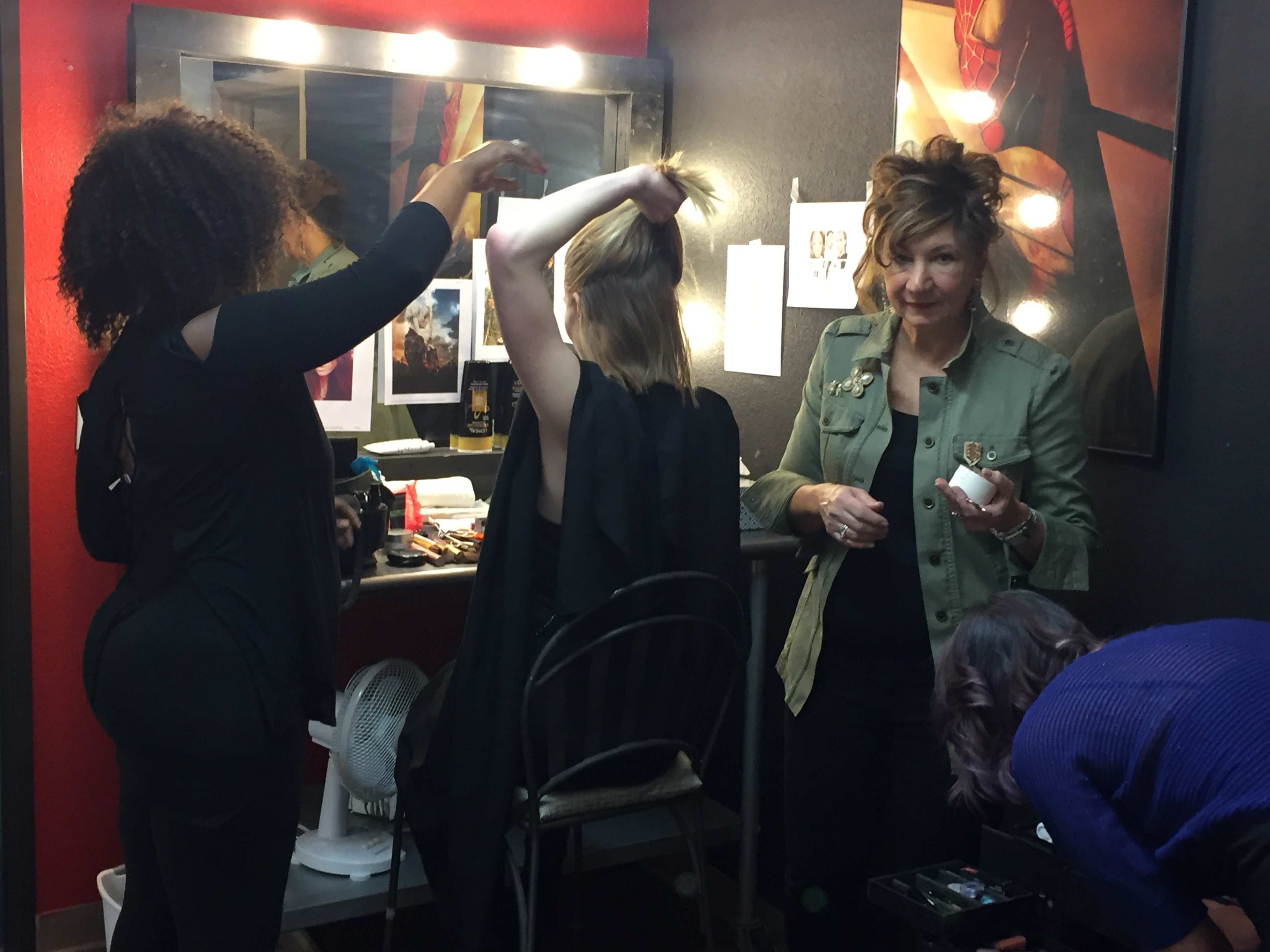 A group of four individuals are situated in a makeup area, with two people styling hair, one applying makeup, and a woman standing nearby observing.
