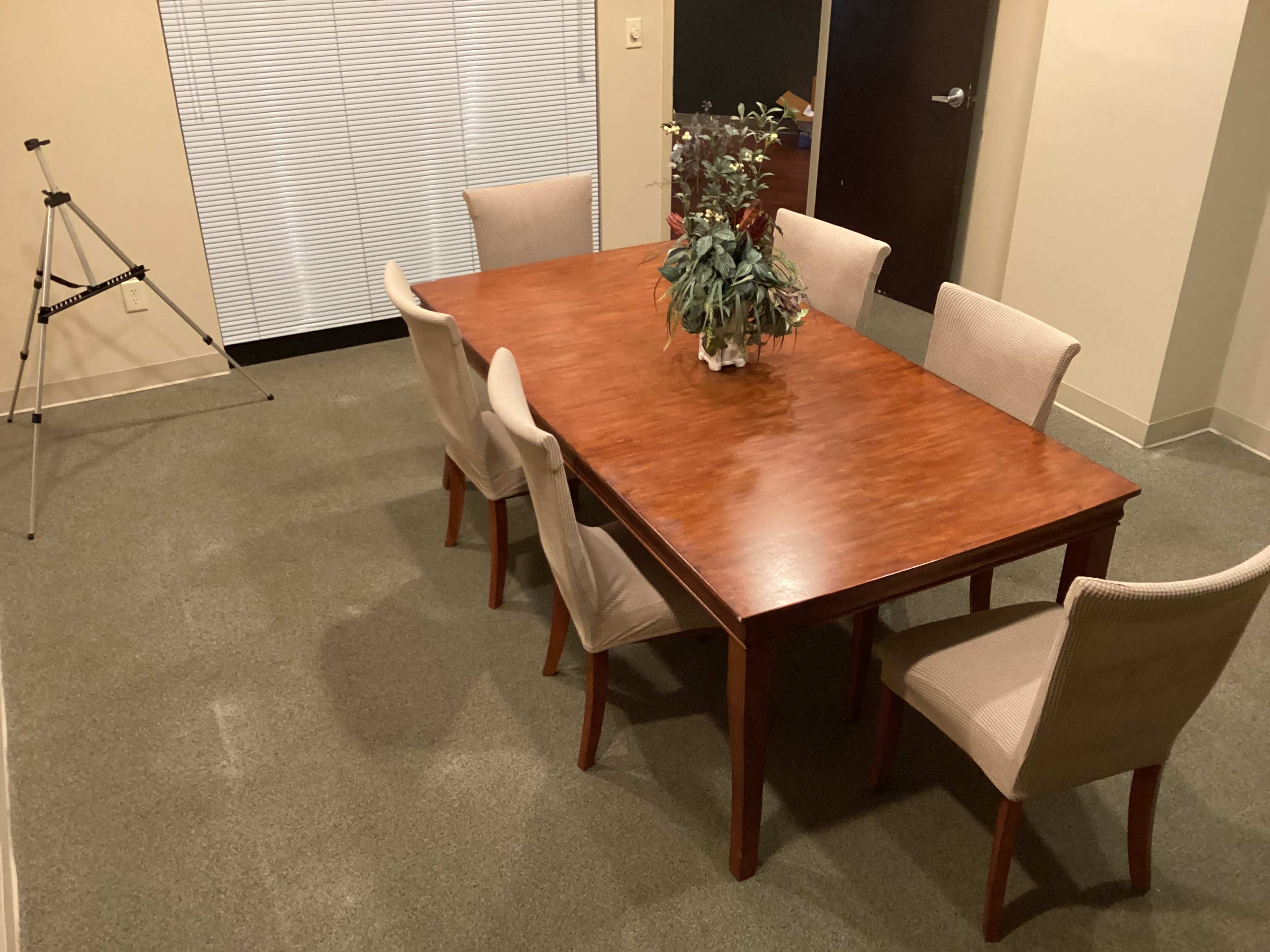 A wooden dining table with a vase of plants is surrounded by six chairs in a room with beige walls and a window covered by blinds.