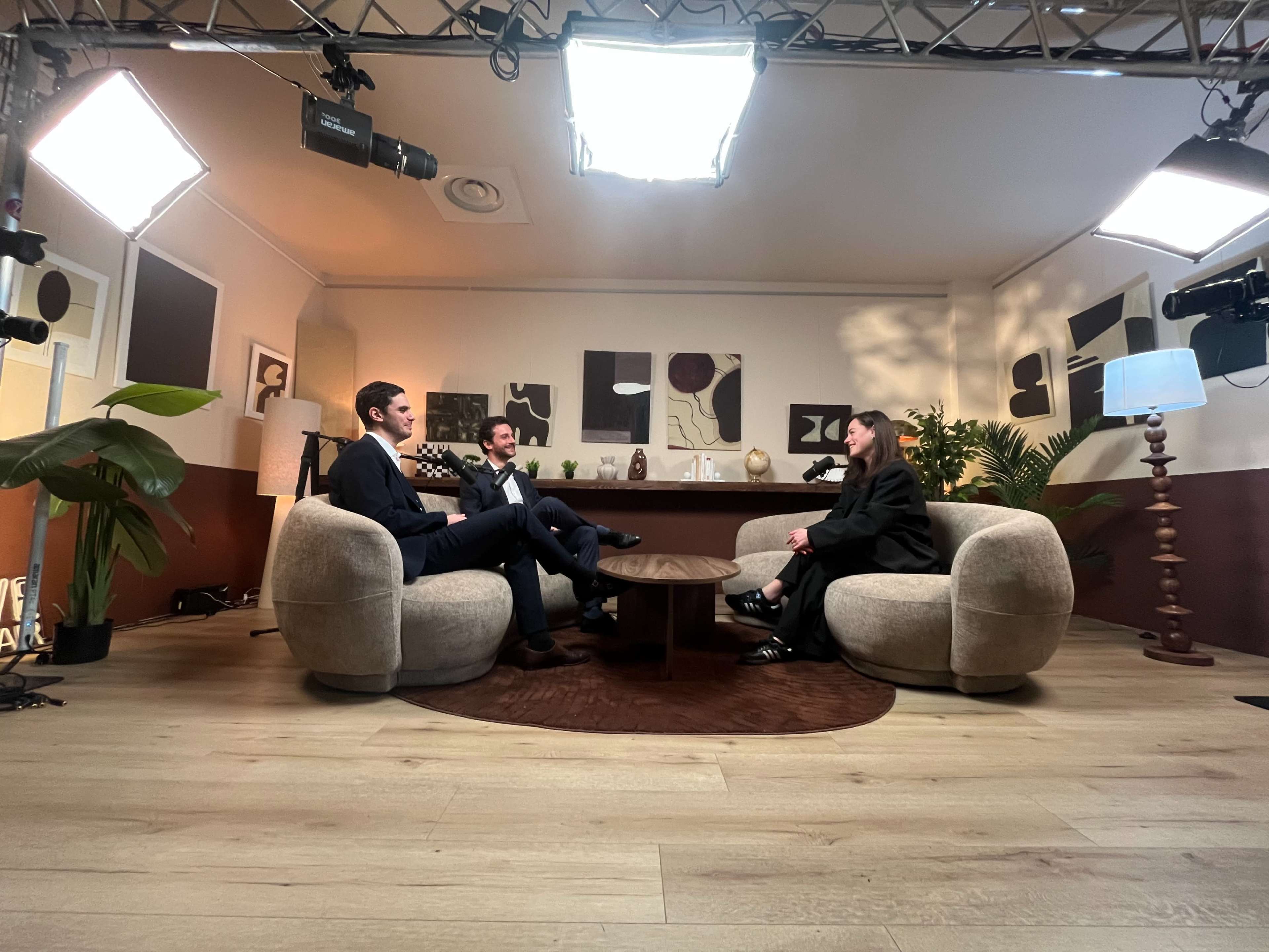 Three people are seated in a modern, well-lit studio with a round coffee table, featuring a large artwork in the background and greenery in the corners.