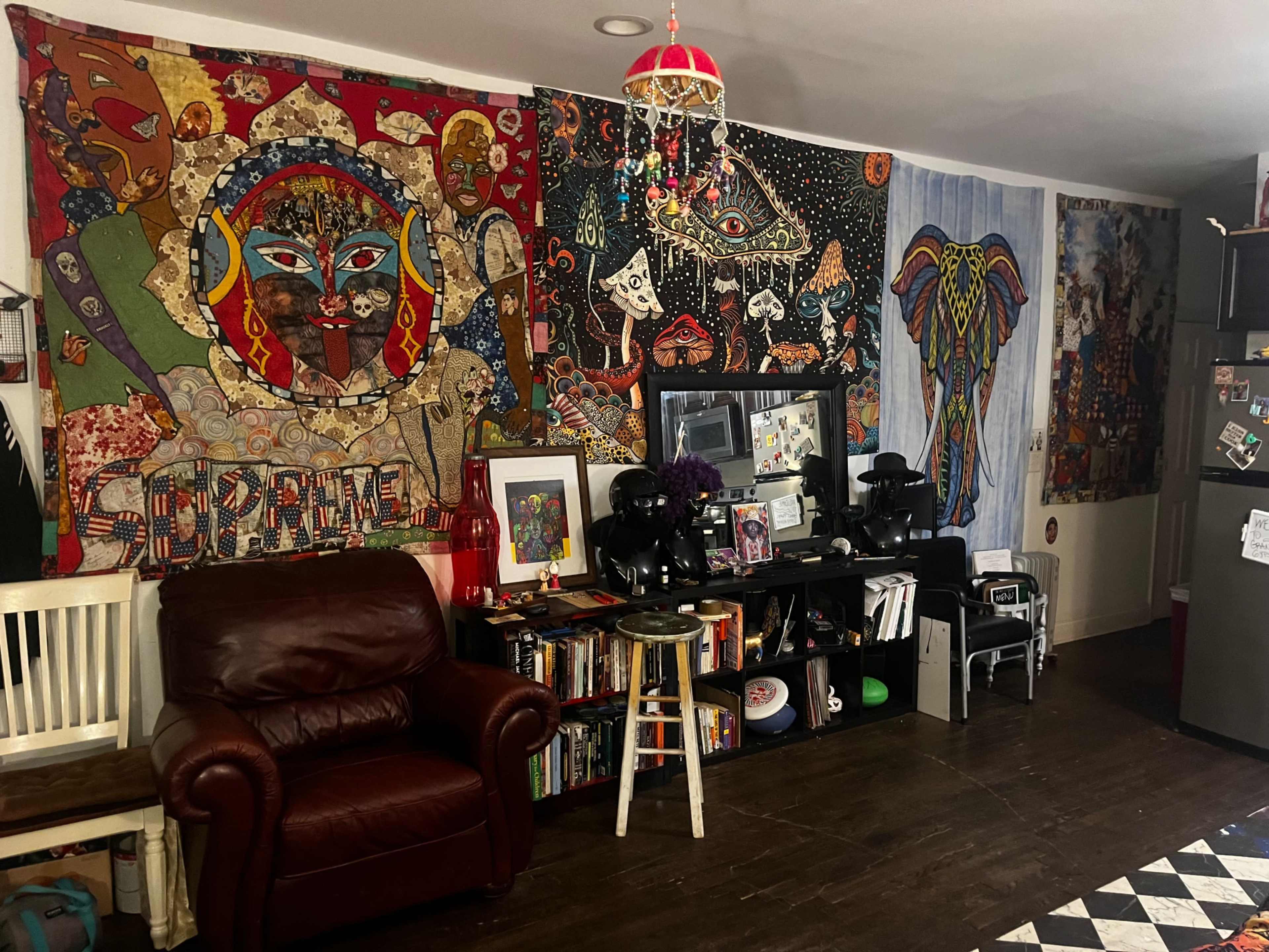 The image shows a cozy living room with colorful wall tapestries, a red leather armchair, a wooden stool, and shelves filled with books and decorative items.