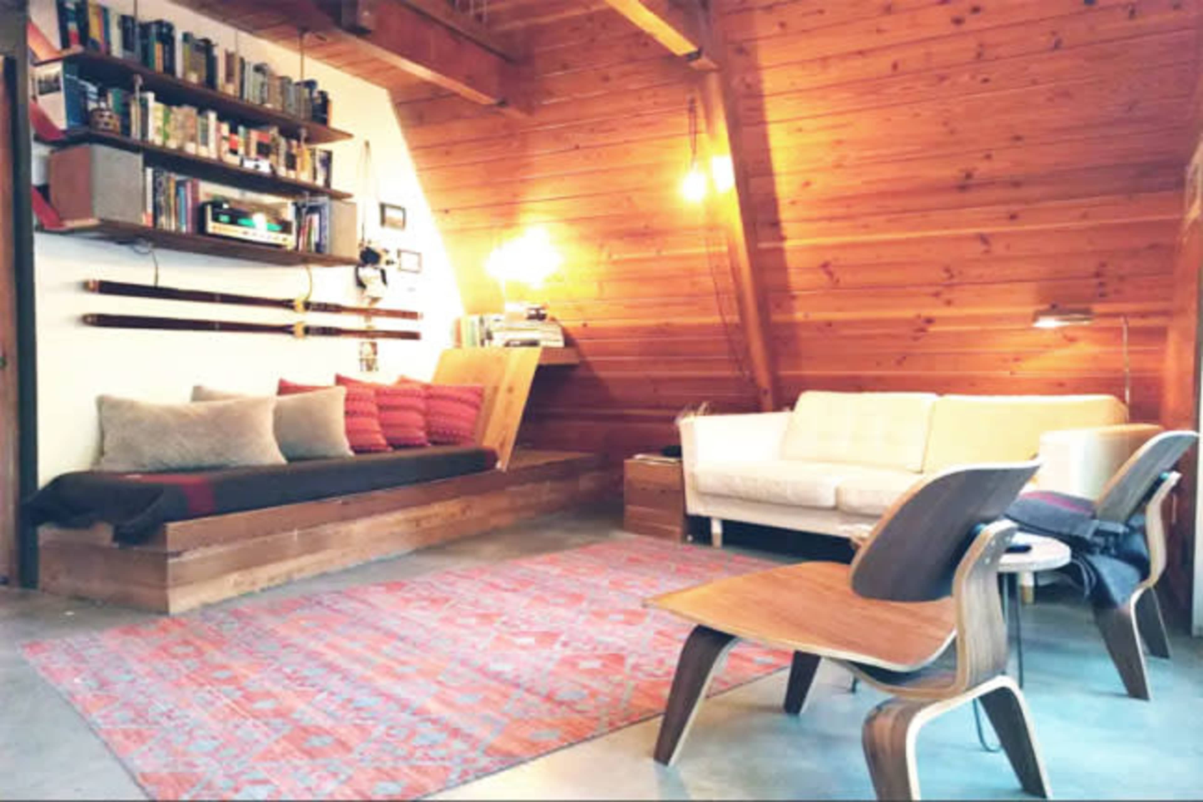 A cozy wooden interior of a living room featuring a built-in seating area with cushions, shelves of books, a white sofa, and a patterned rug.