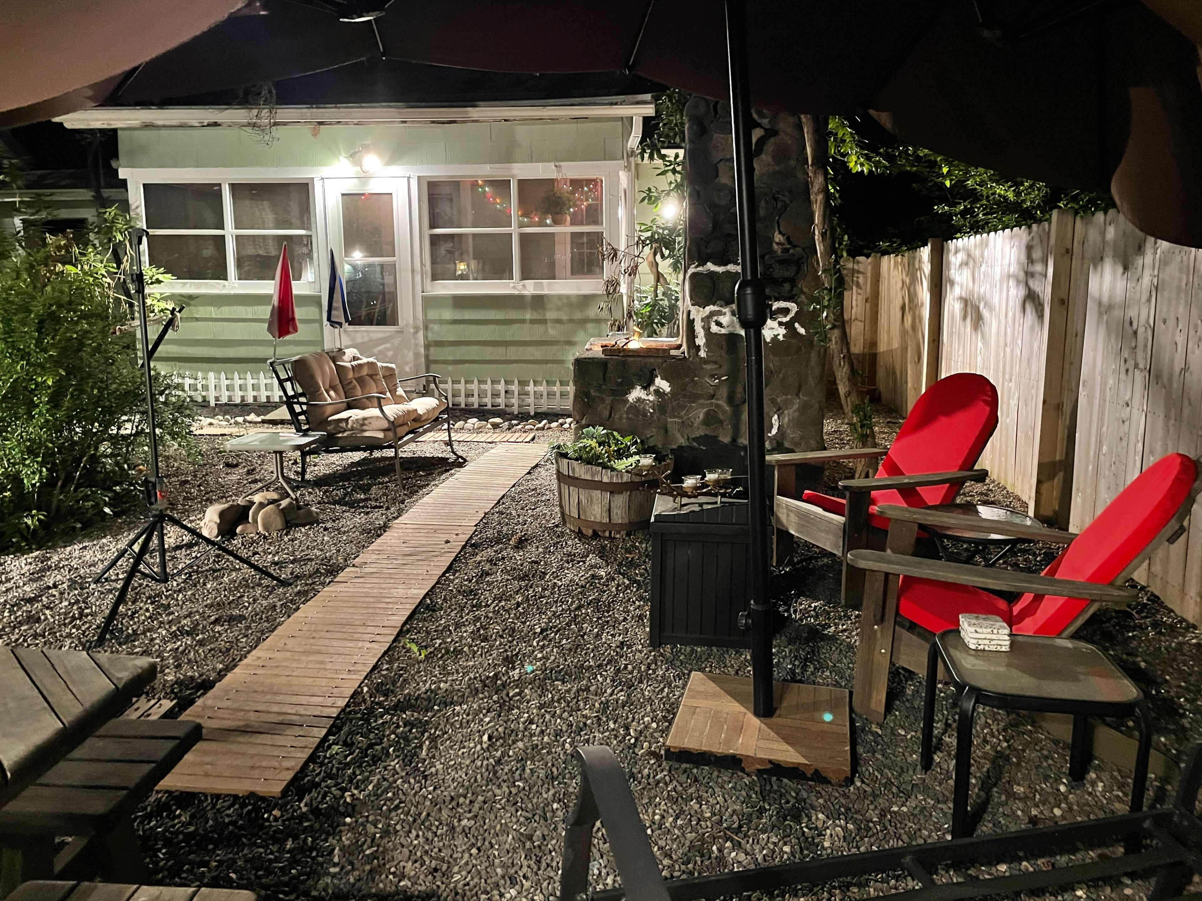 The image shows a cozy outdoor garden space featuring two red chairs, a wooden pathway, and a small seating area beside a house with a lit window.