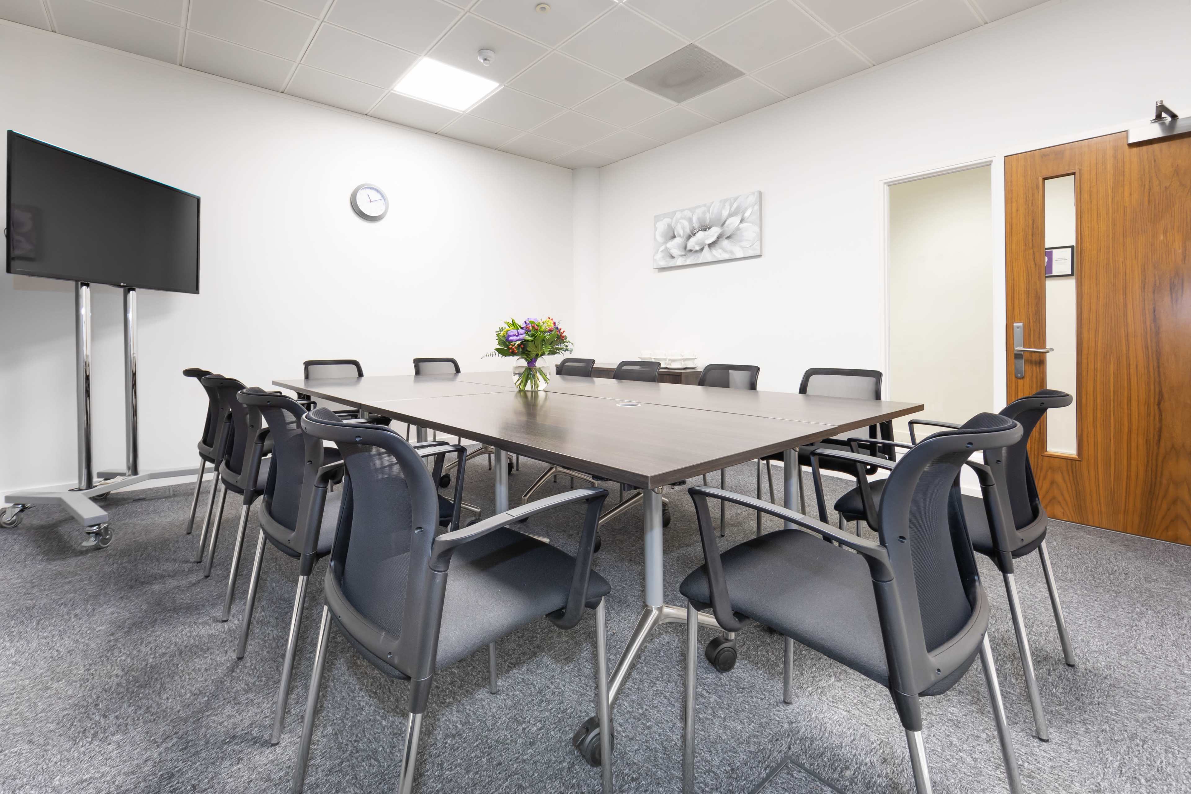 A large, rectangular table surrounded by ten black chairs is positioned in a well-lit conference room with a TV on a stand and a floral arrangement in the center.