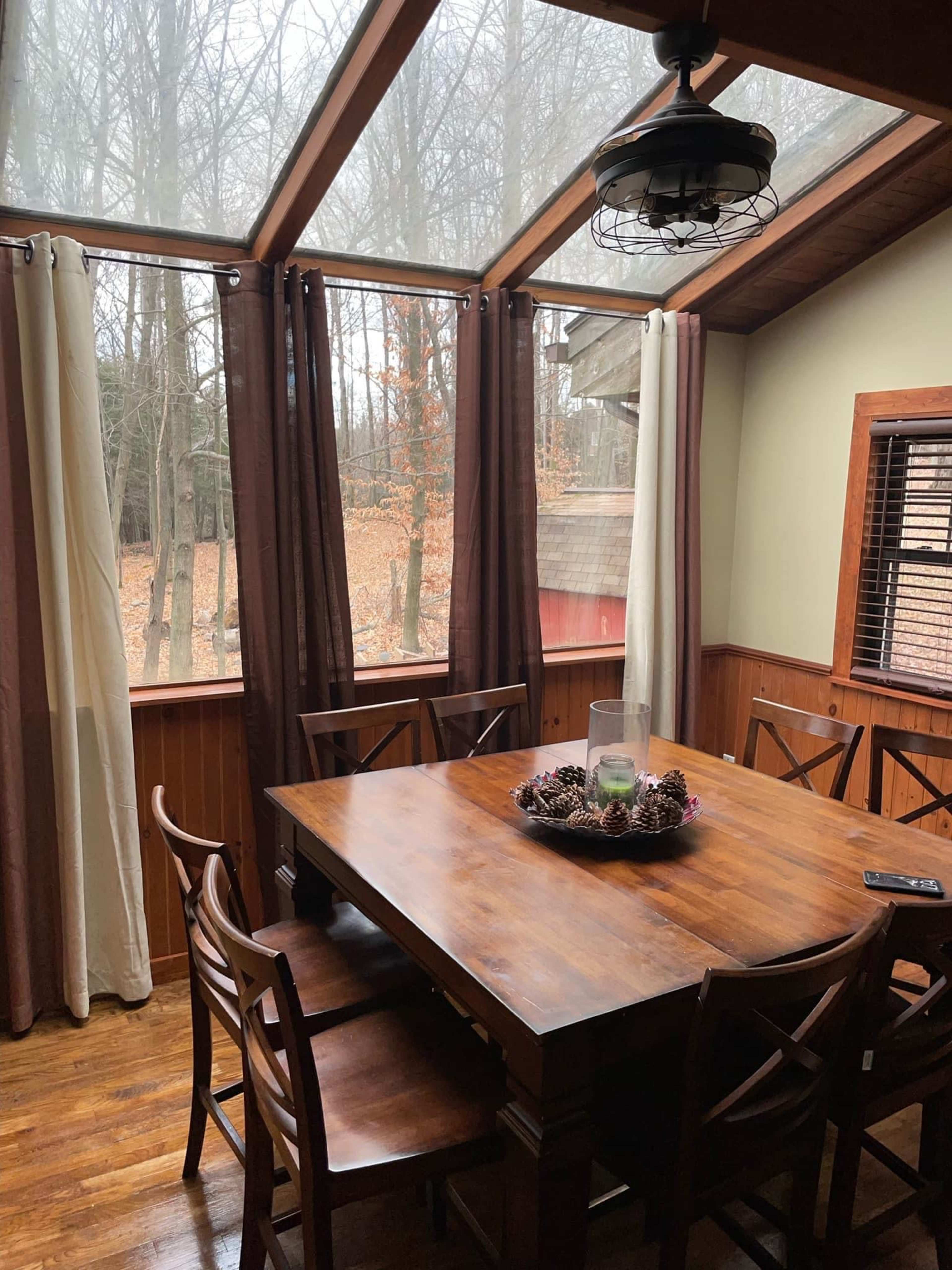 A wooden dining table surrounded by chairs is positioned in a sunlit room with large windows overlooking a wooded area.