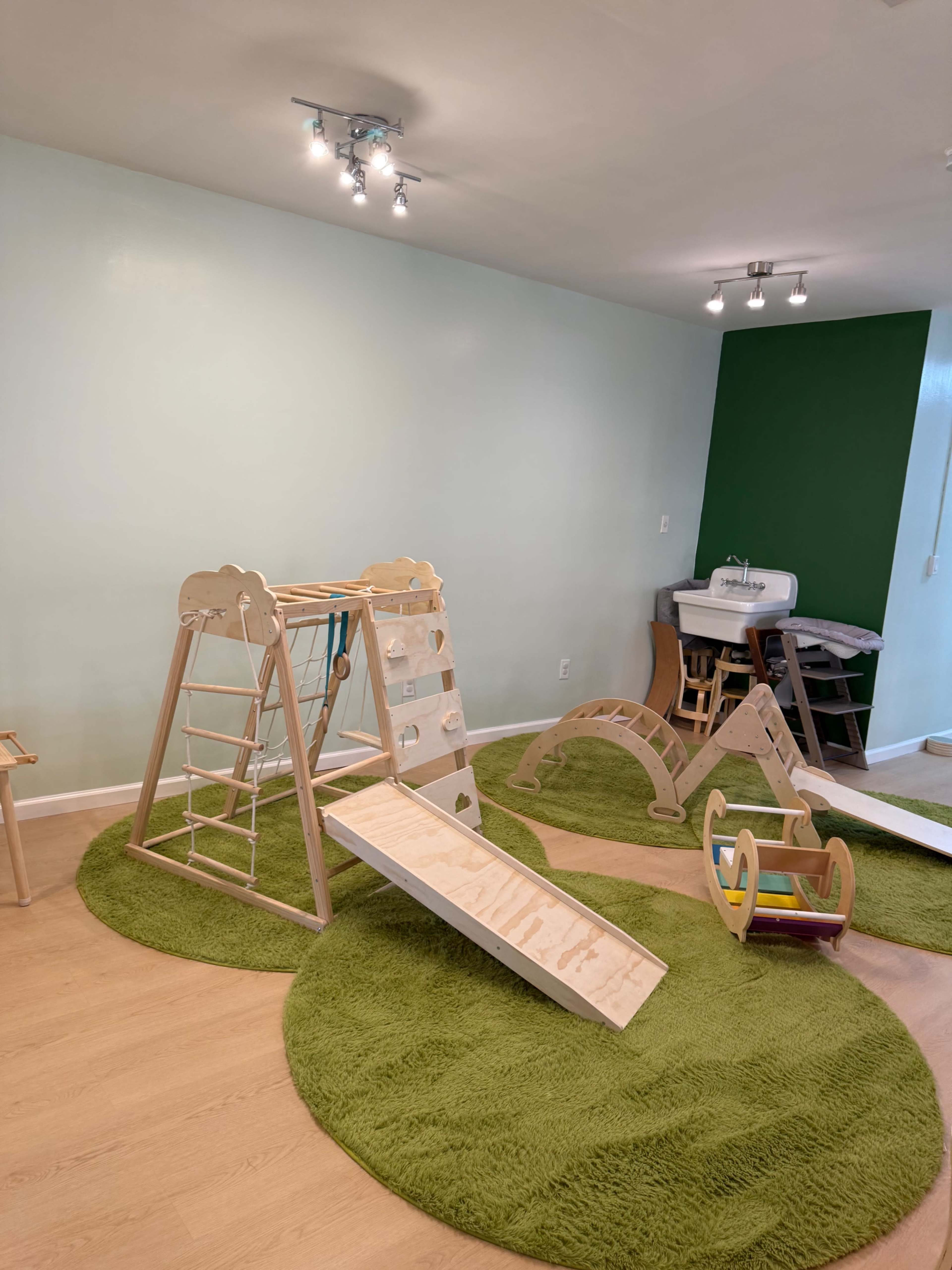 The image shows a playroom with wooden play equipment, a green carpet, and a sink area in the corner.