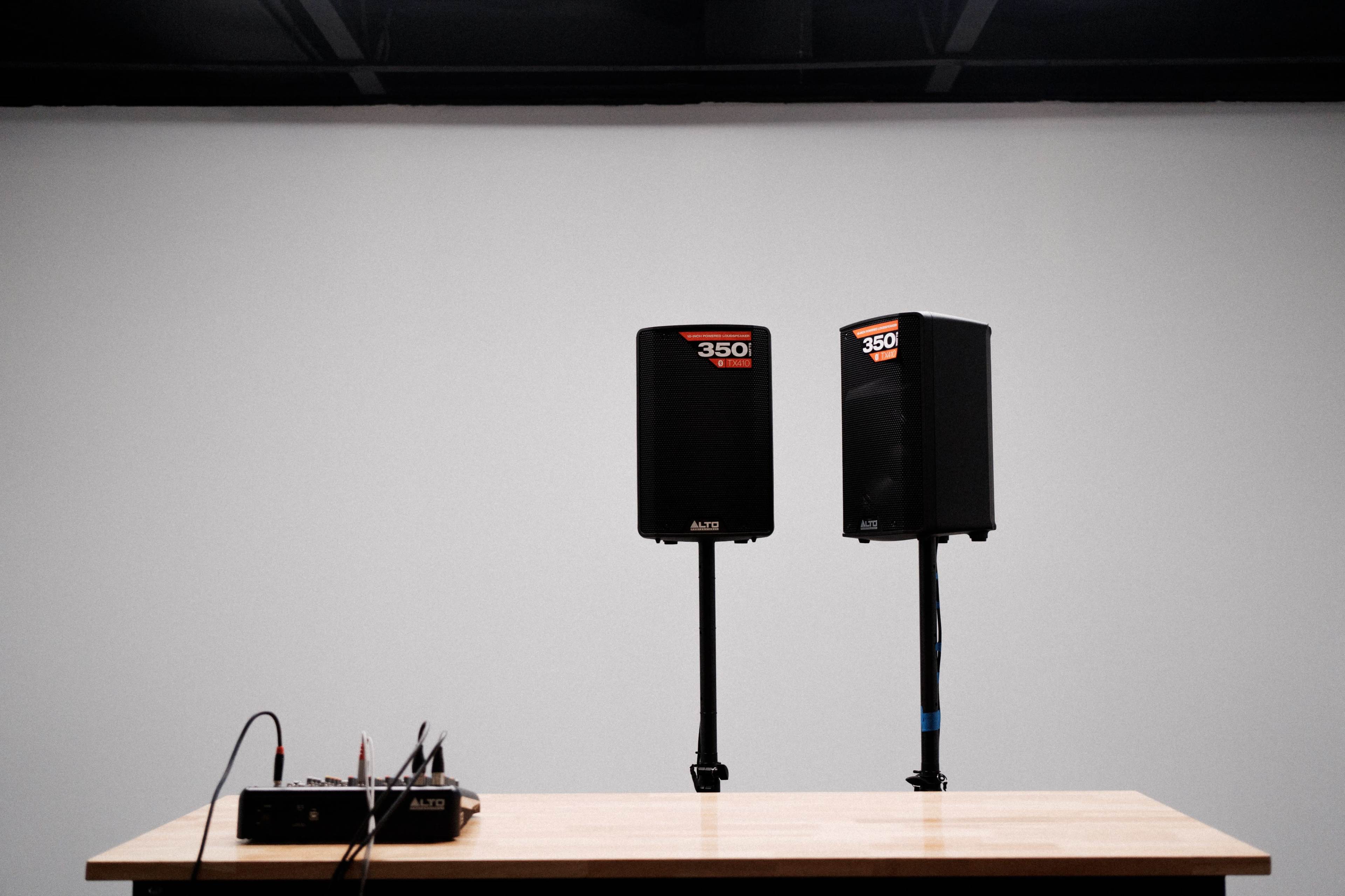 Two speaker systems are positioned on stands beside an audio mixer on a wooden table against a plain white wall.