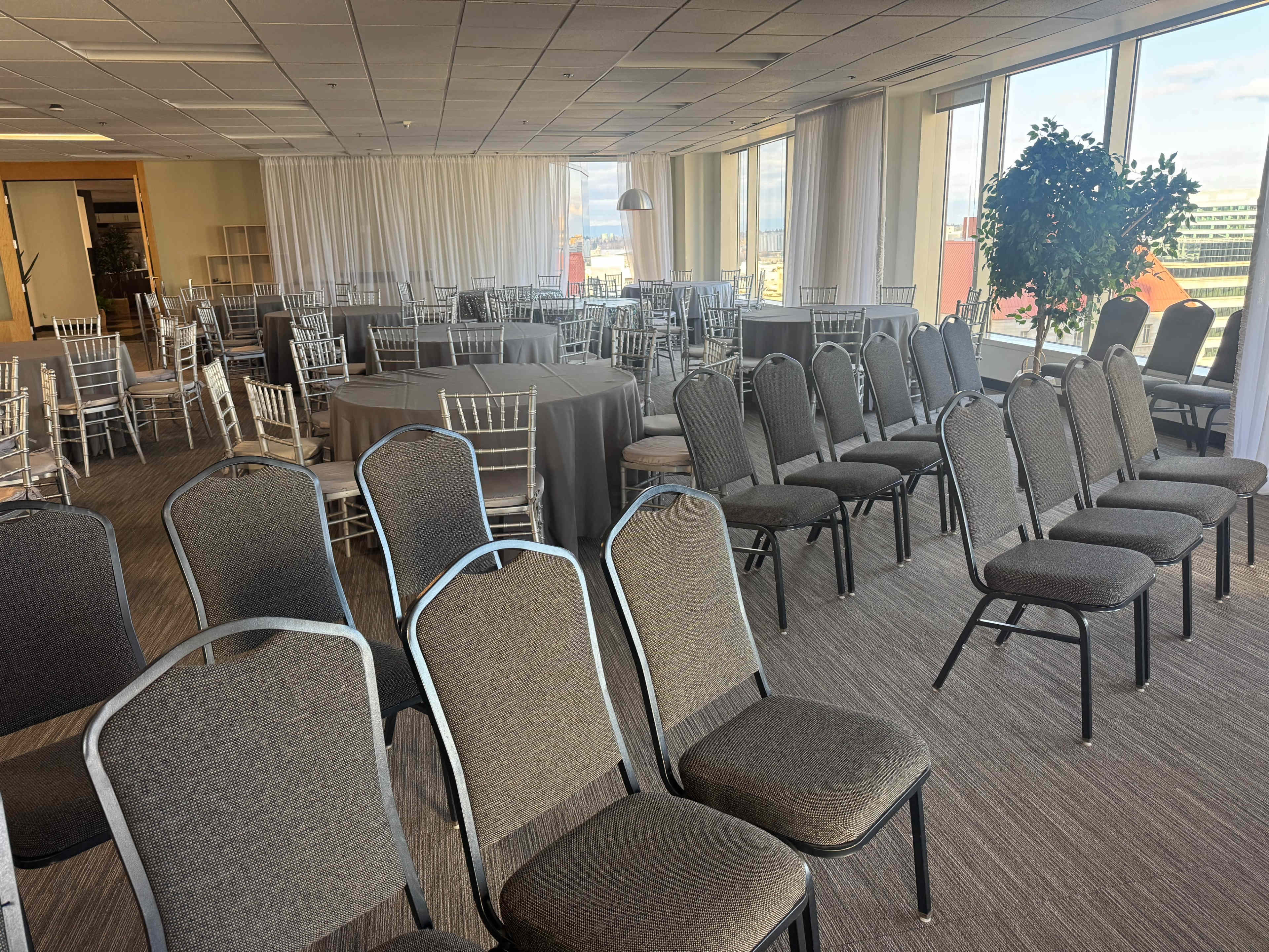 The image shows a well-lit event space featuring rows of chairs and tables arranged for a gathering, with large windows offering a view outside.