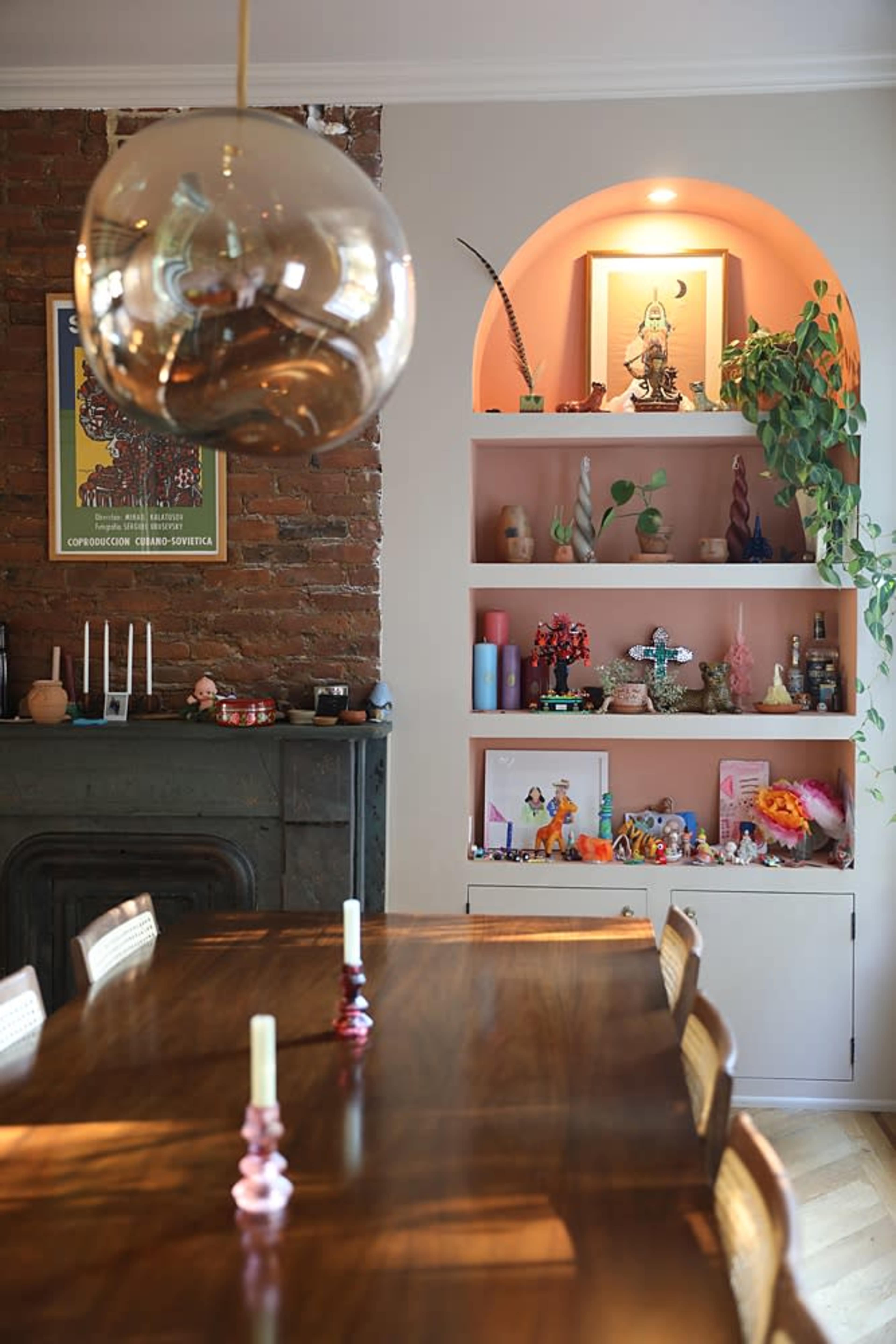 The image shows a dining area with a long wooden table, candles at the center, and a wall featuring shelves filled with decorative items and a small statue.