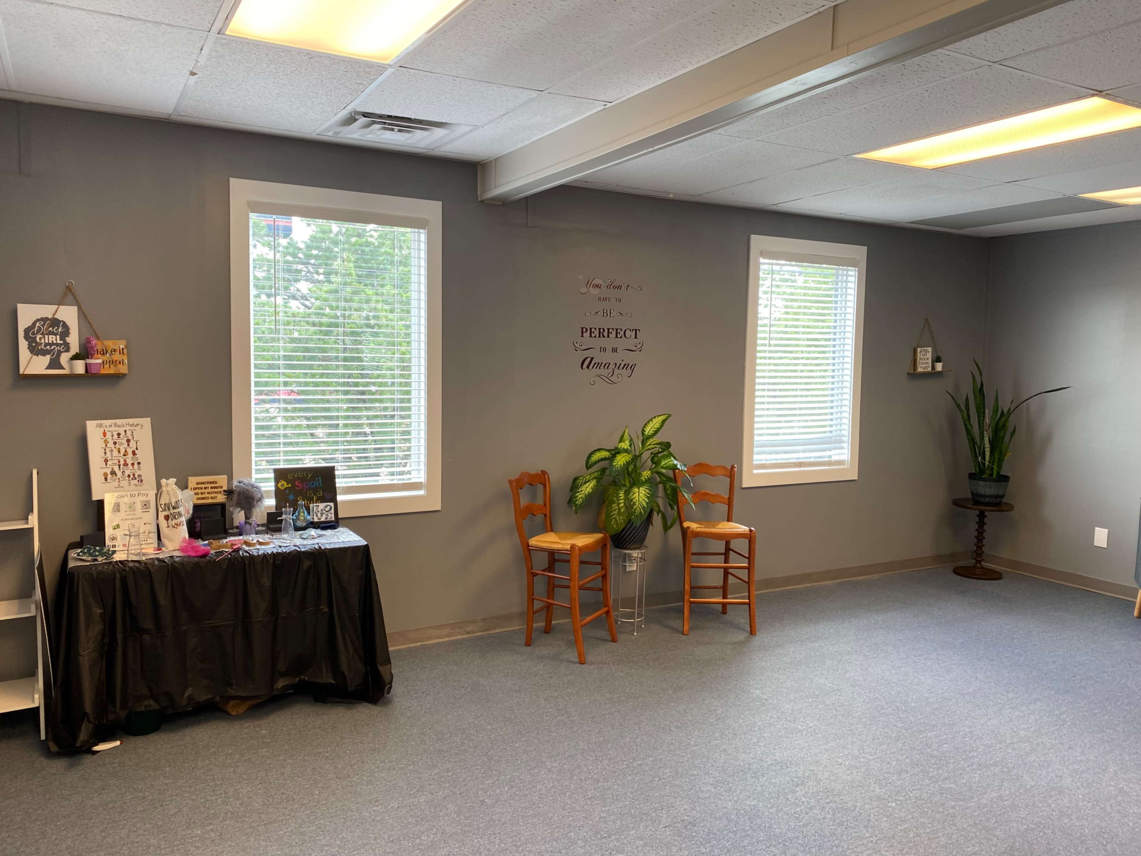 A sparsely furnished room with gray walls, featuring two wooden chairs, a plant, and a black table displaying various items against one wall.