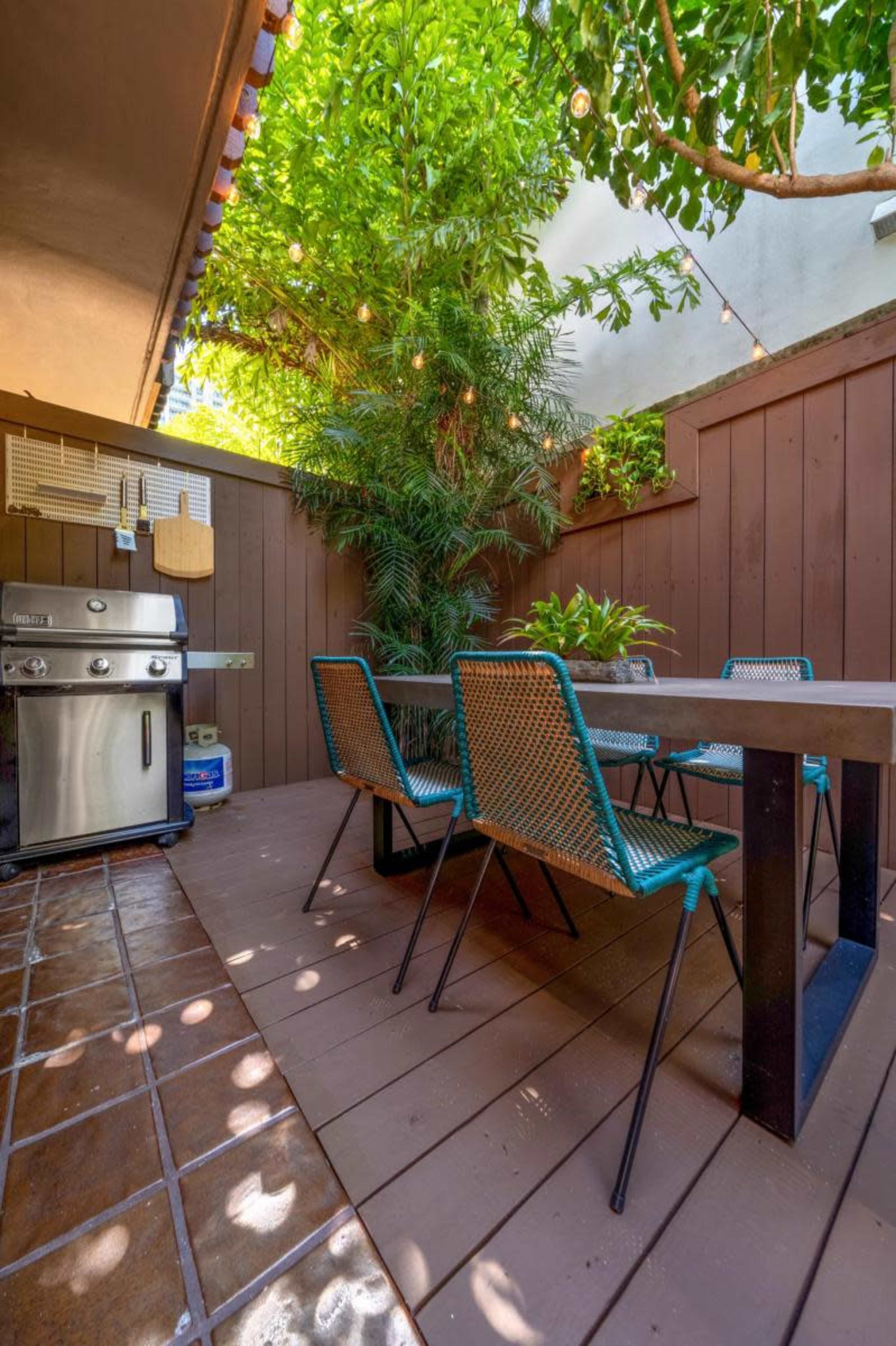 A small, enclosed outdoor patio features a dining table surrounded by chairs, a grill, and lush green plants.