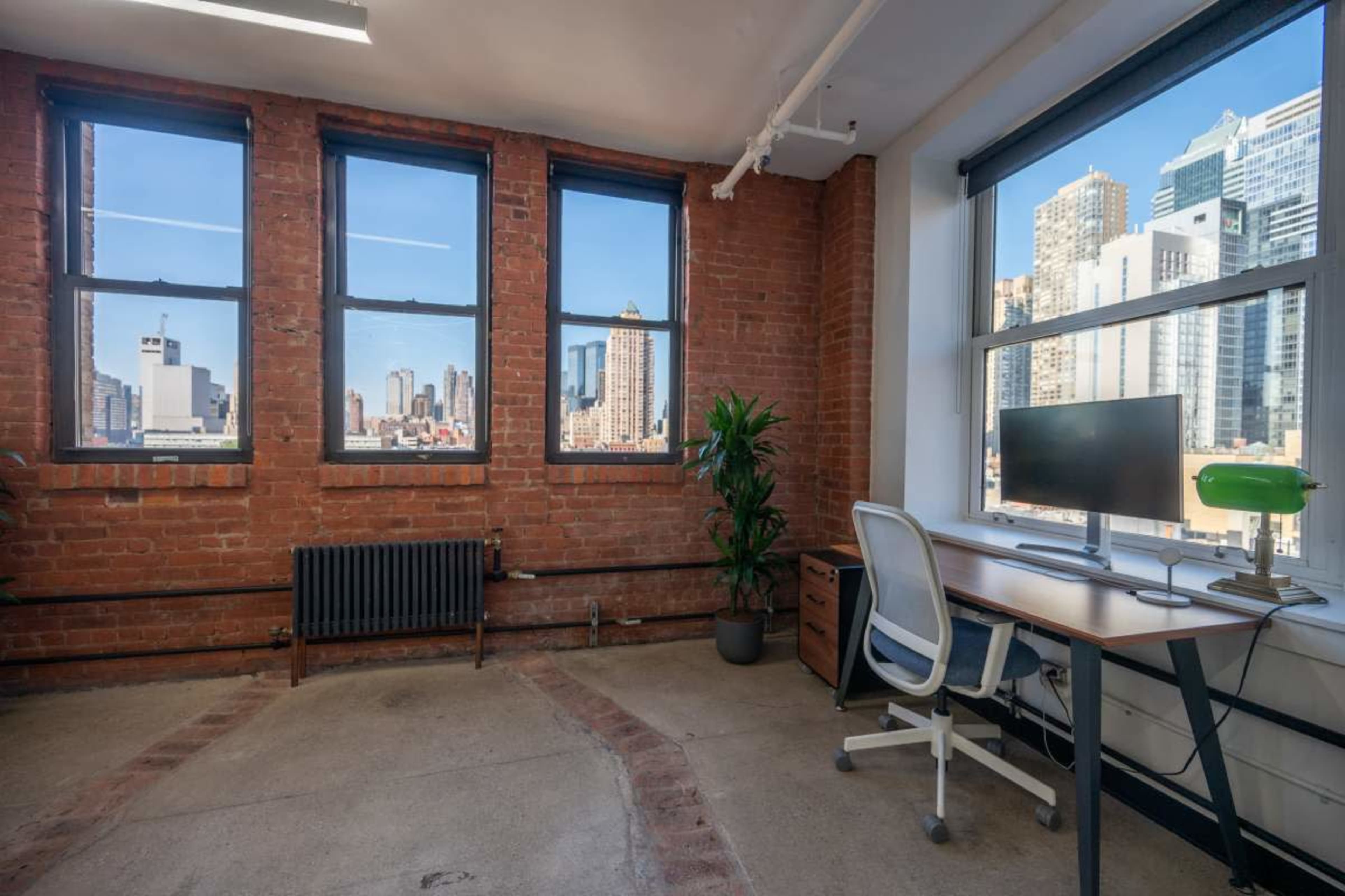 The image shows a minimalist office space featuring a desk with a monitor and a green lamp, large windows revealing a city skyline, and exposed brick walls.