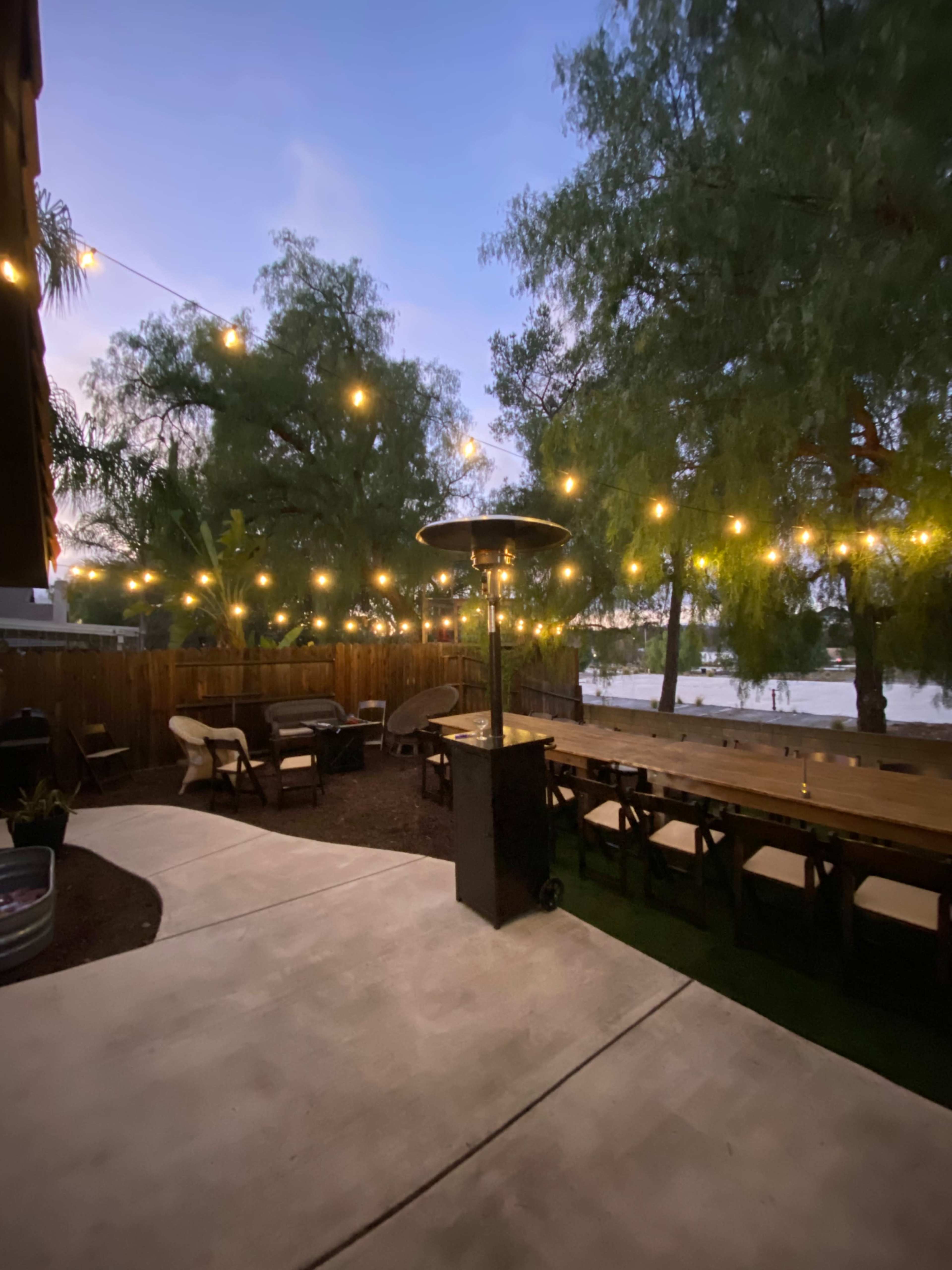 The image shows a patio area with a long dining table, string lights overhead, and a heater, overlooking a water view.