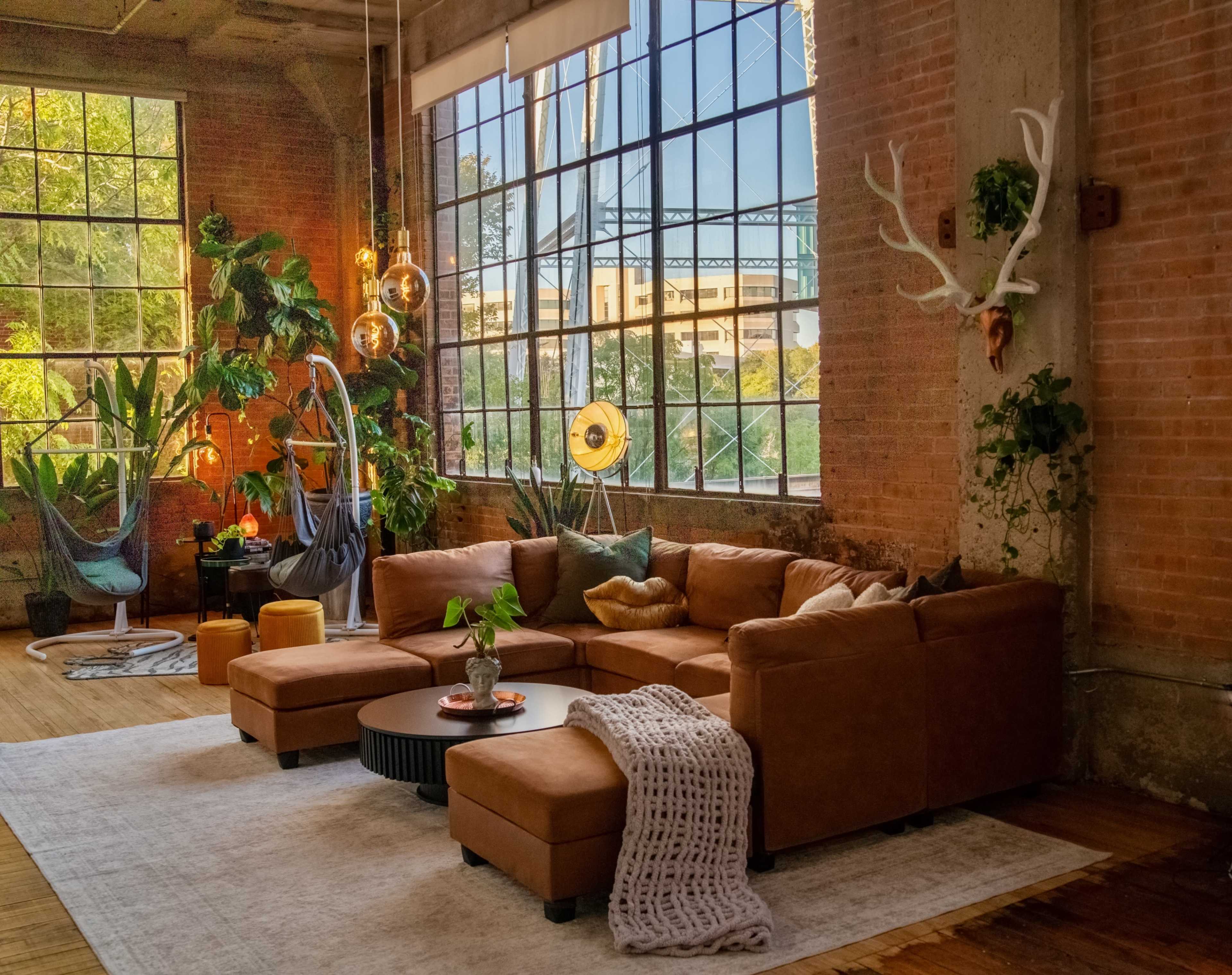 The image shows a spacious industrial-style living room with large windows, a brown sectional sofa, plants, and decorative lighting.
