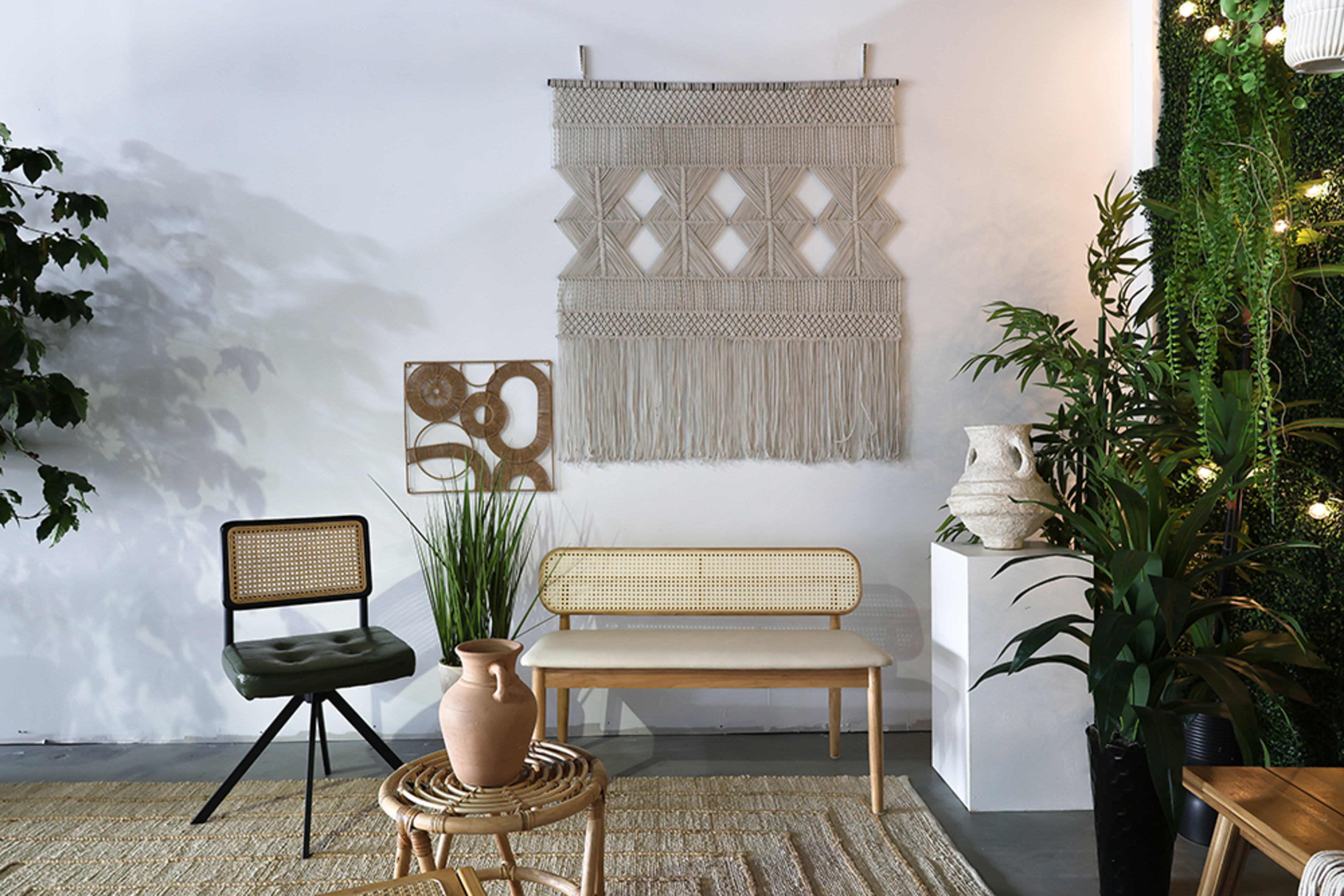 The image shows a modern interior space featuring a macramé wall hanging, a rattan bench, a chair with a green cushion, a decorative pot, and various plants.
