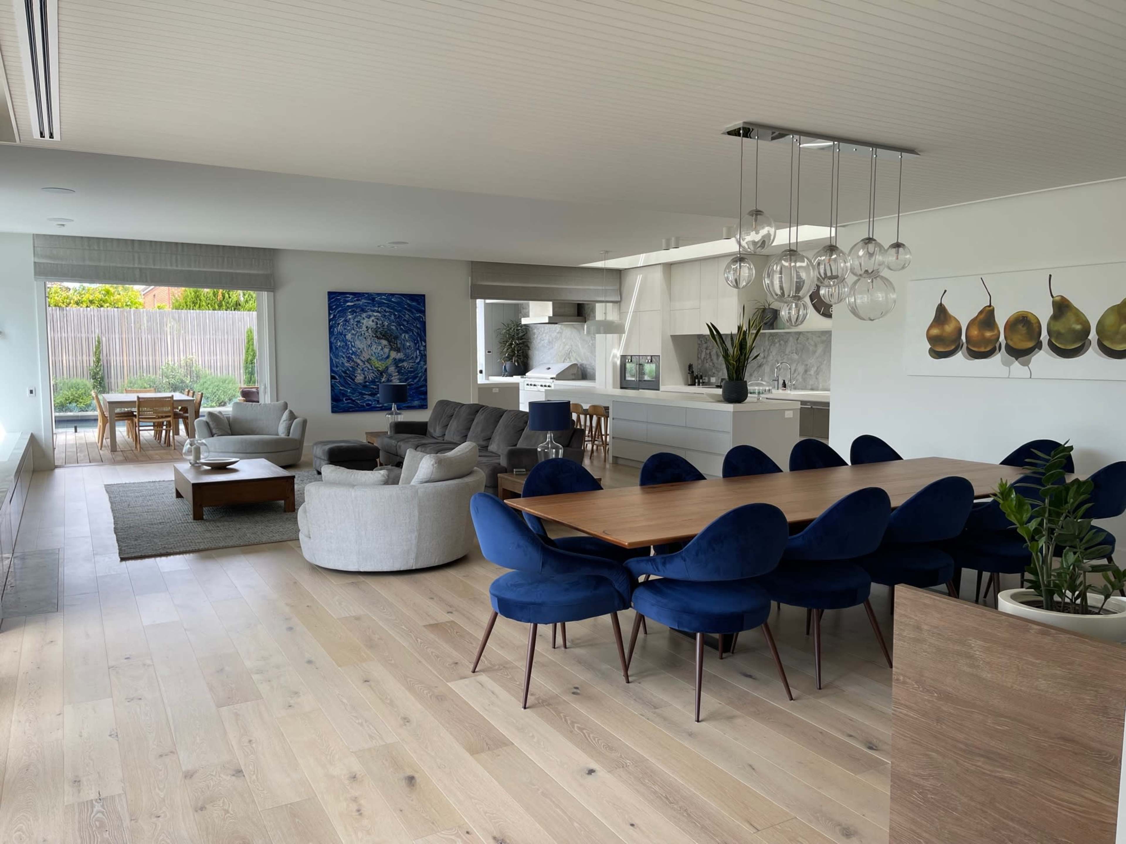 A modern open-concept living and dining area featuring a long wooden table surrounded by blue chairs, a gray sectional sofa, and large windows with a view of an outdoor space.