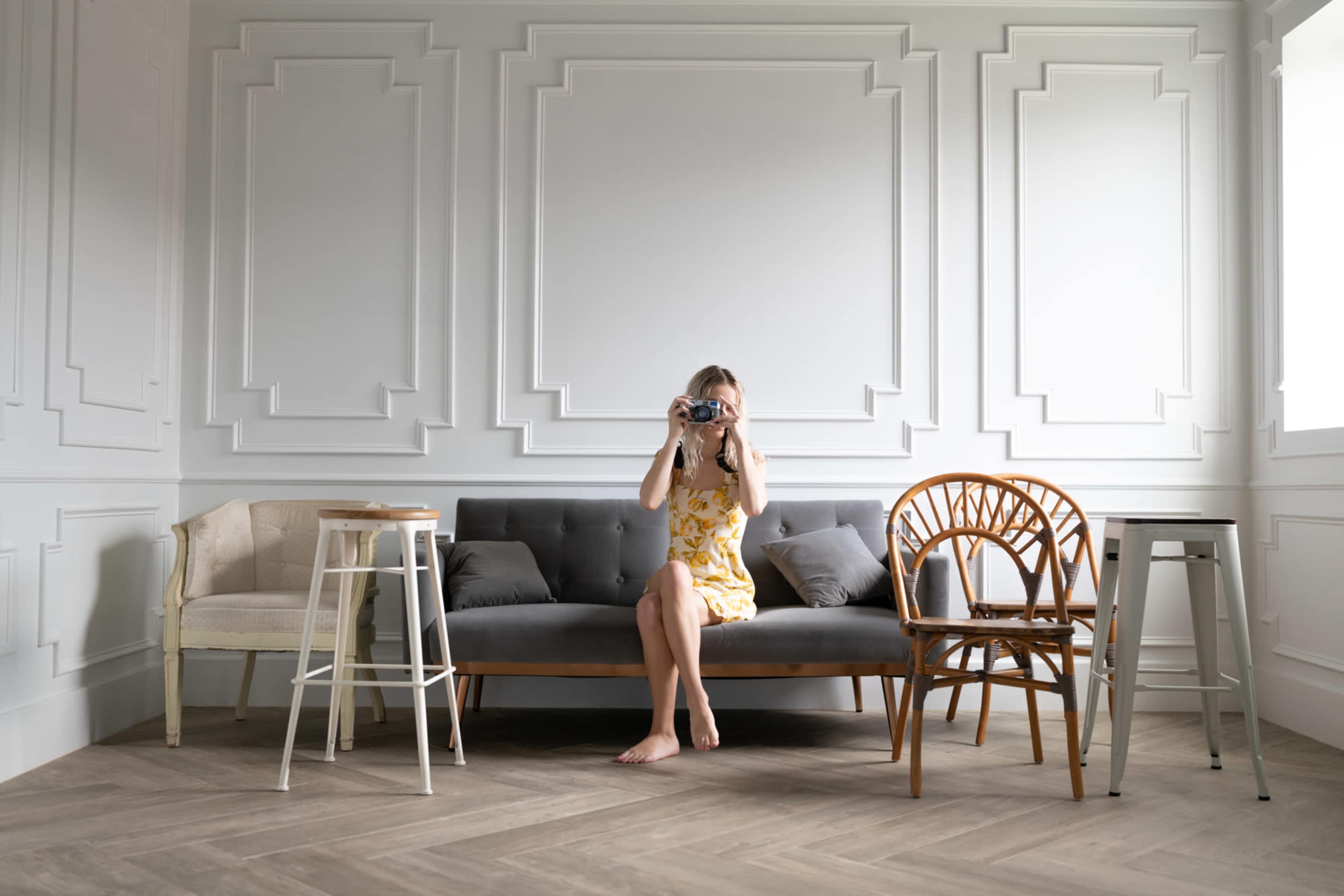 A woman in a yellow dress sits on a gray sofa, holding a camera, in a room with paneled walls and wooden flooring.