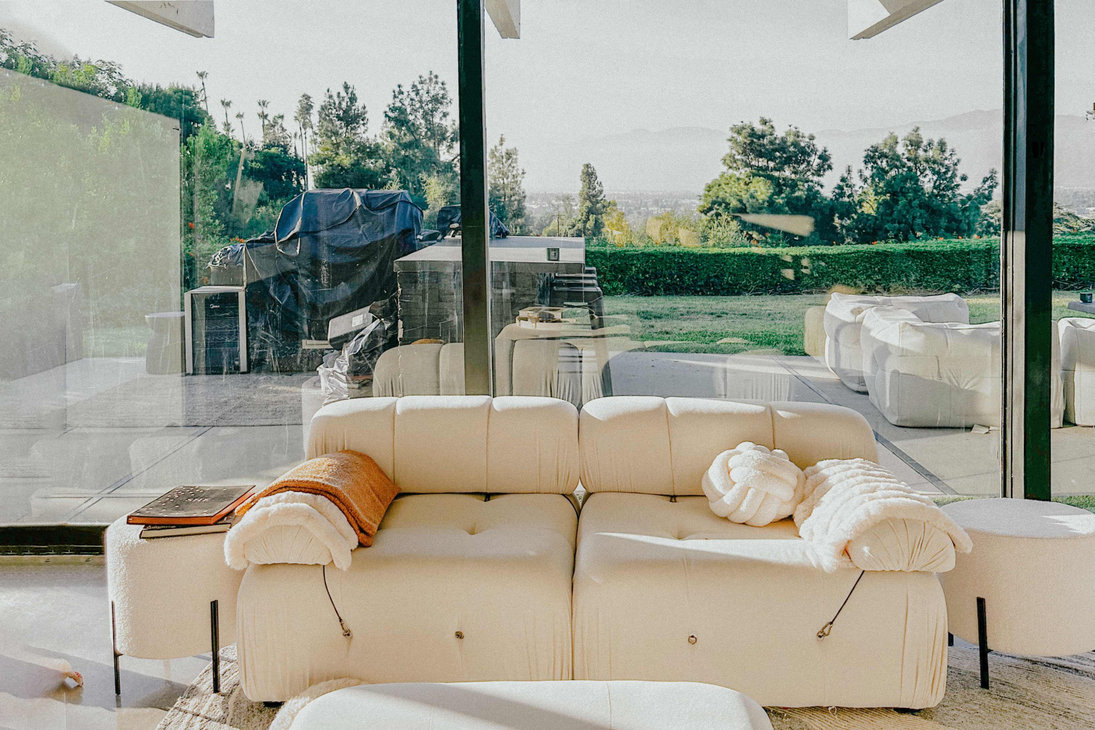 A modern living room features a white couch with soft throws, large glass windows offering a view of a landscaped yard and distant mountains.
