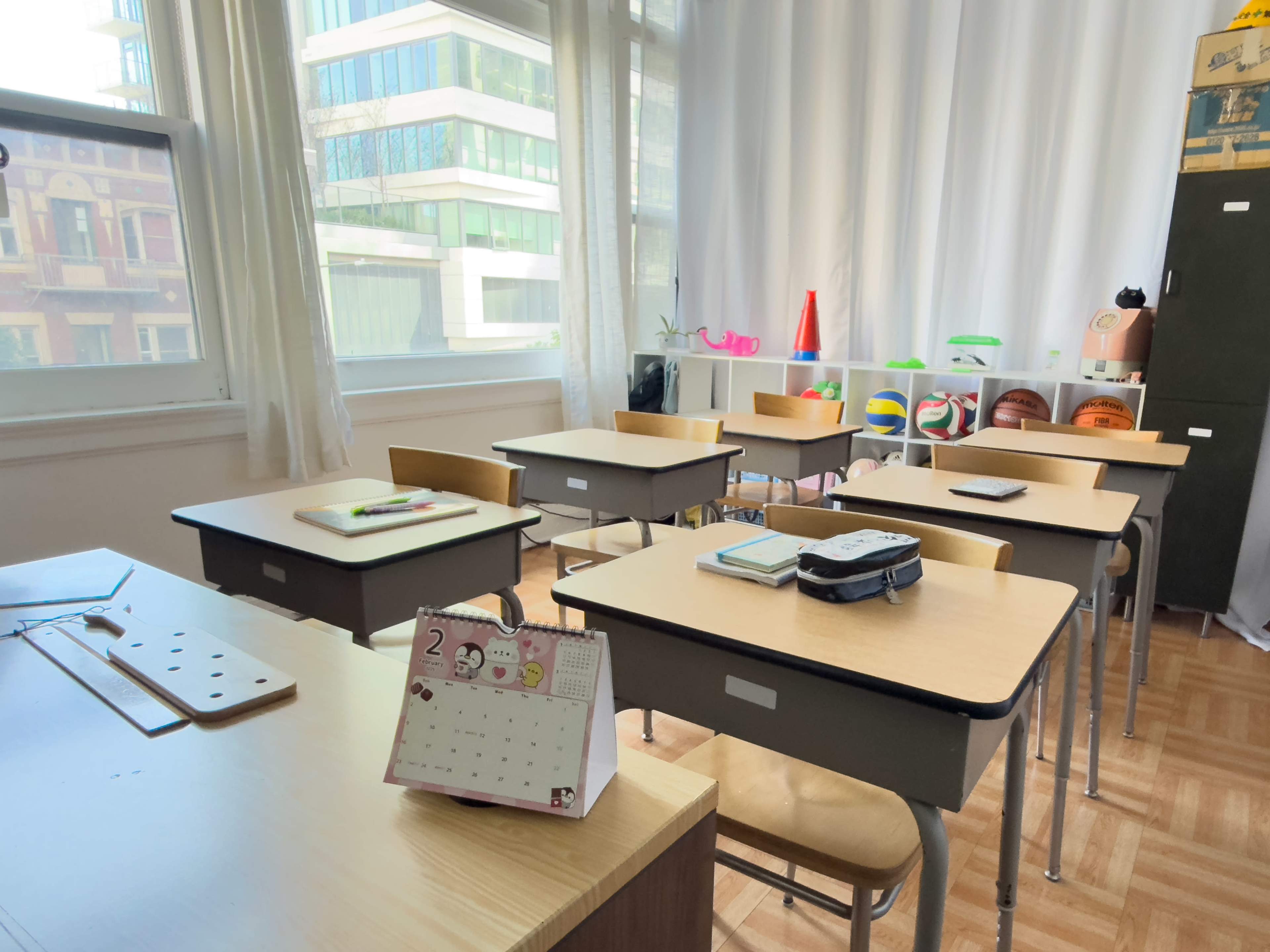 The image shows a bright classroom with several empty desks, a calendar on the foreground, and various educational supplies arranged on shelves.