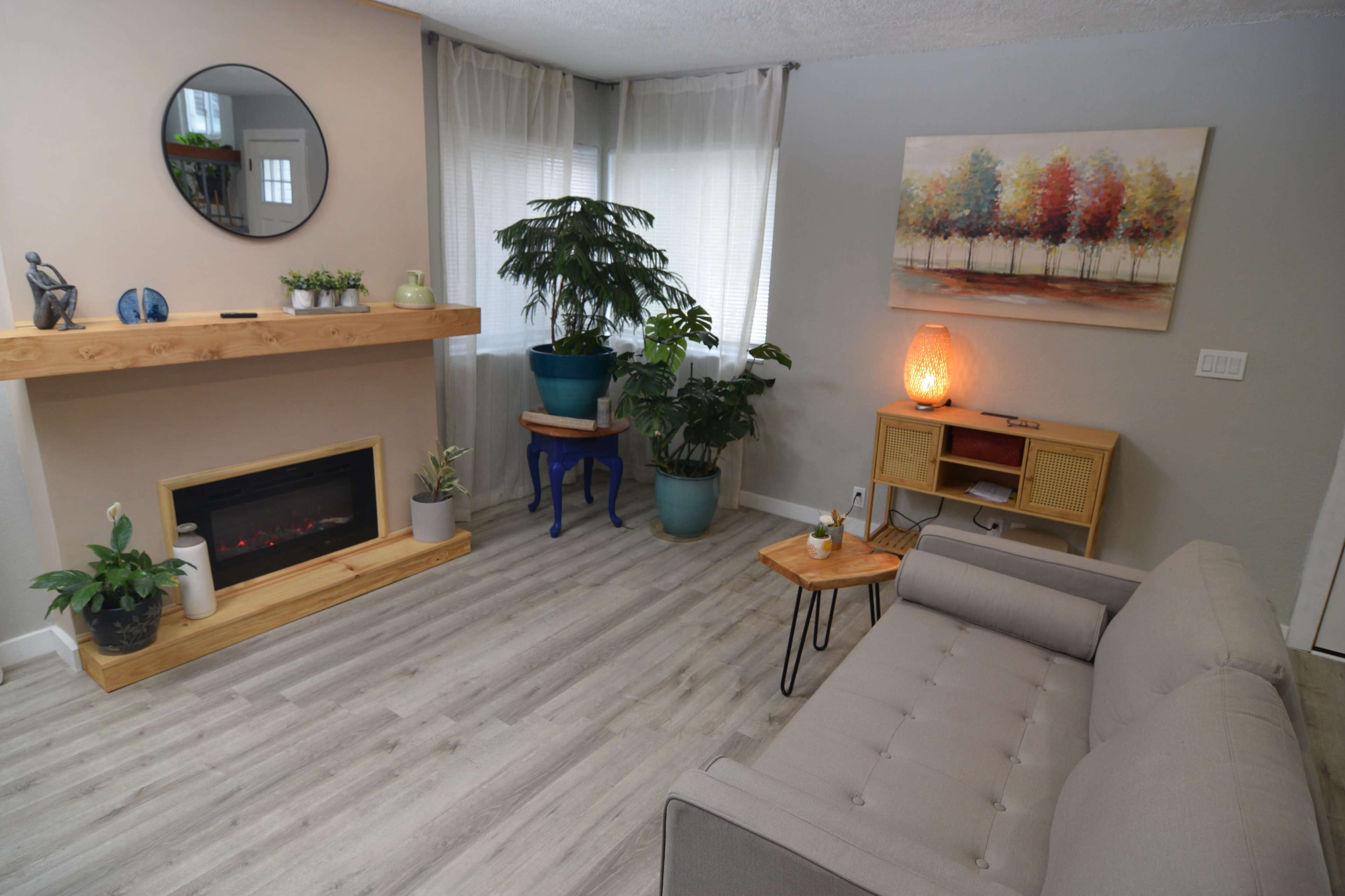 A cozy living room with a gray sofa, a fireplace, a large plant, and decorative artwork on the walls.