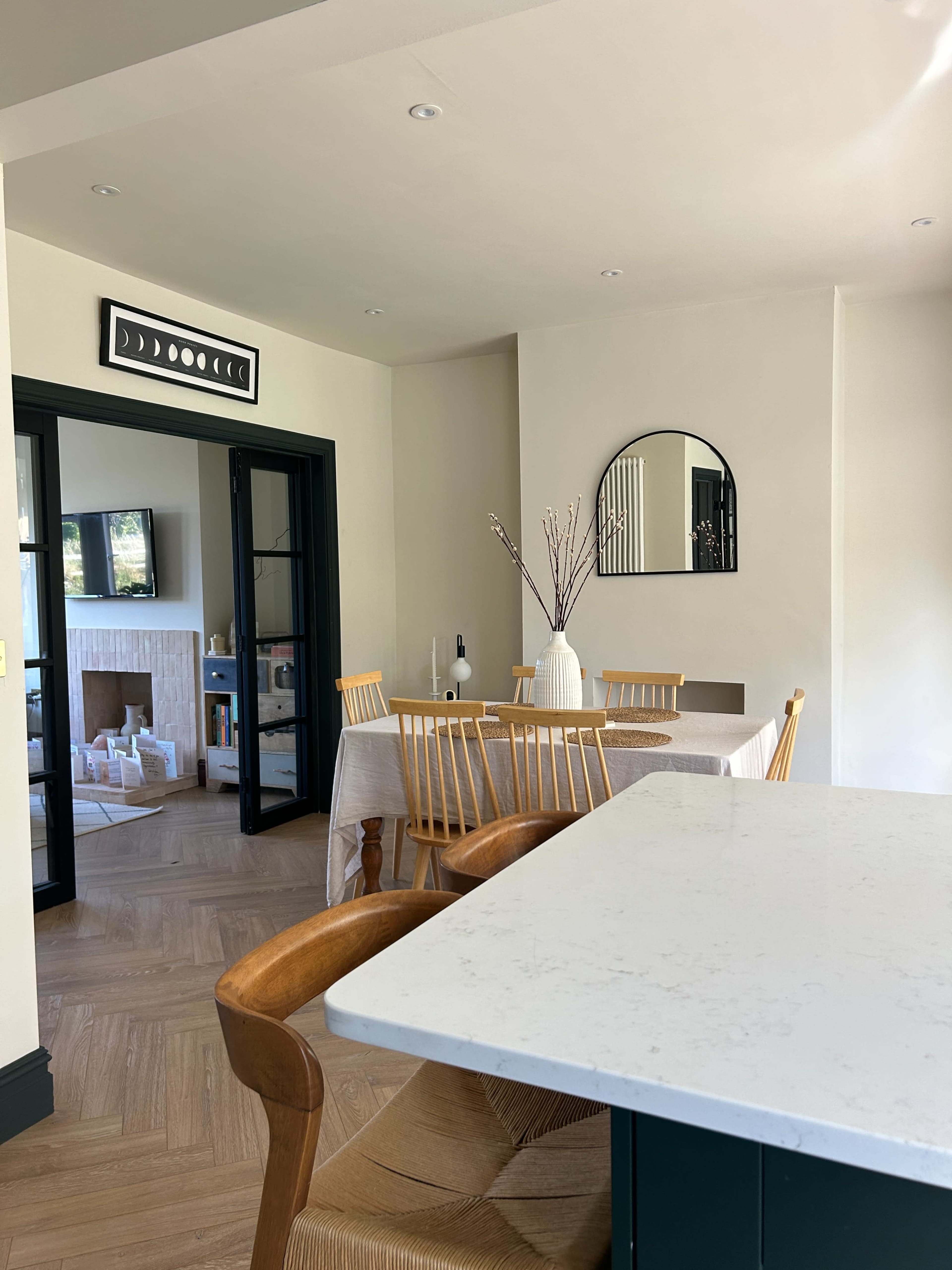 A dining area features a light-colored table with wooden chairs, surrounded by minimal decor and an adjacent living space visible through glass doors.