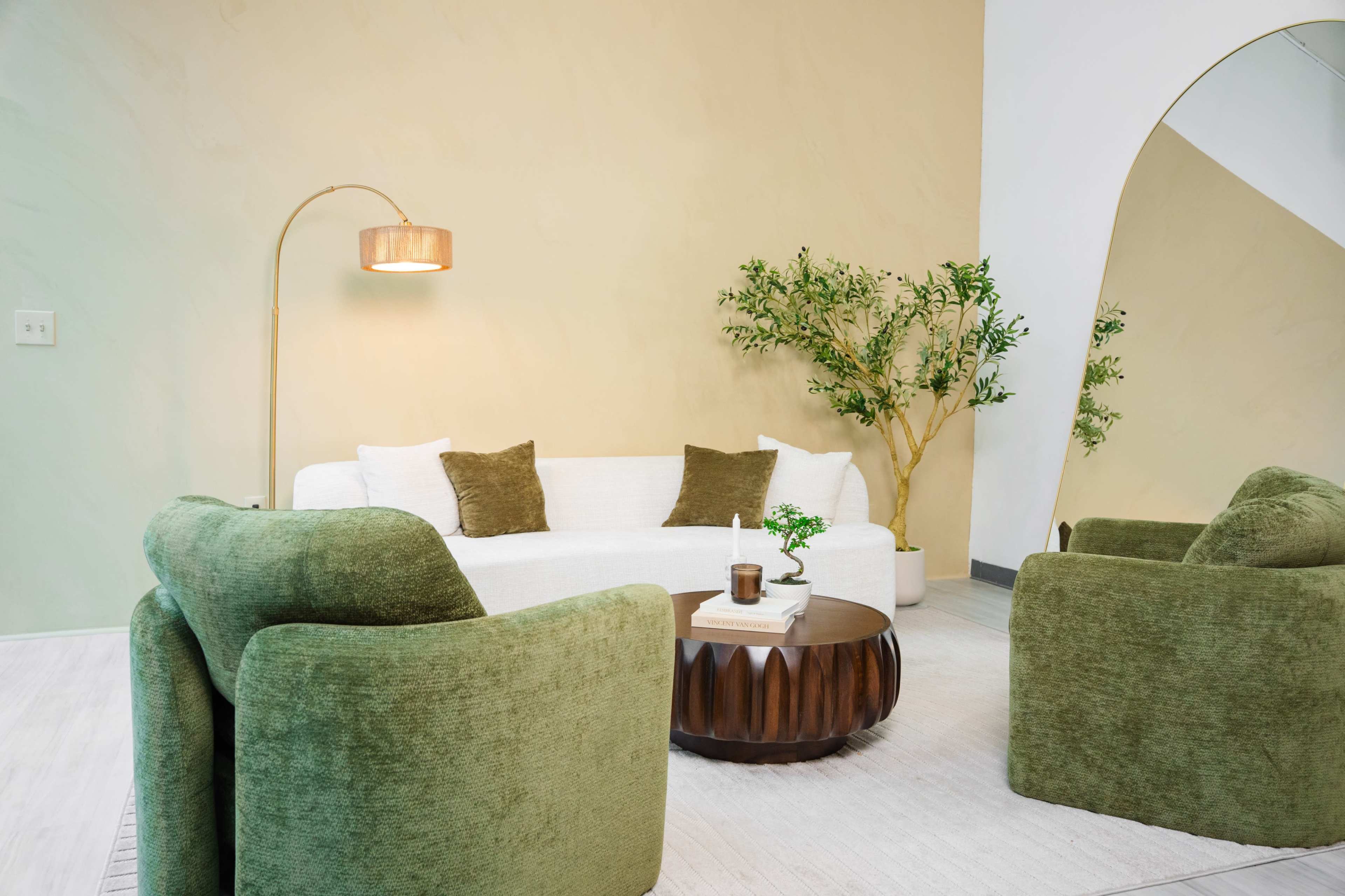 The image shows a modern living room featuring a white sofa, two green armchairs, a round wooden coffee table, a floor lamp, and a decorative plant against a neutral wall.