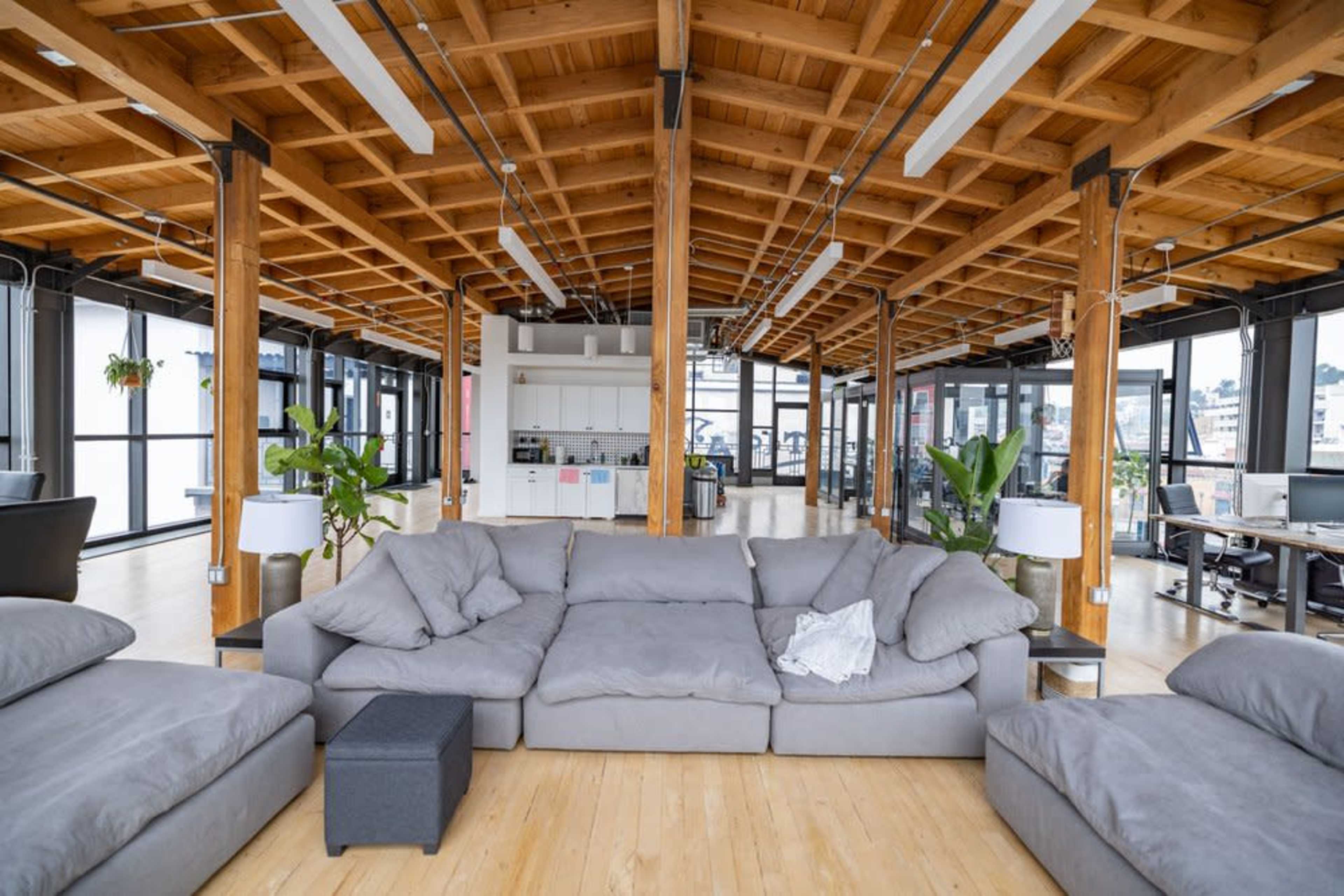 A spacious, modern office interior with a large, gray sectional sofa in the foreground and large windows providing natural light.
