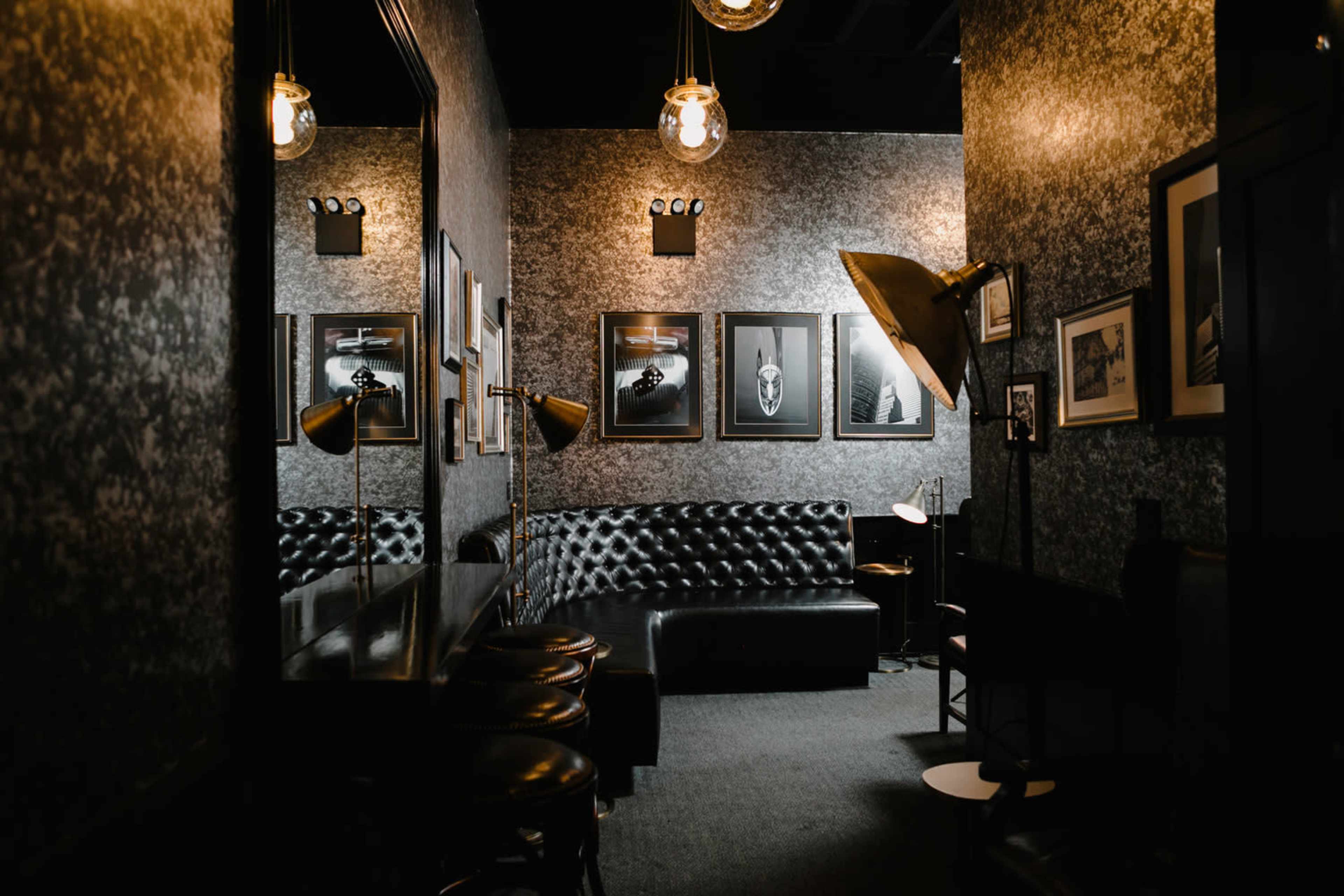 A dimly lit lounge with dark walls, a tufted black leather sofa, and framed black-and-white photographs.