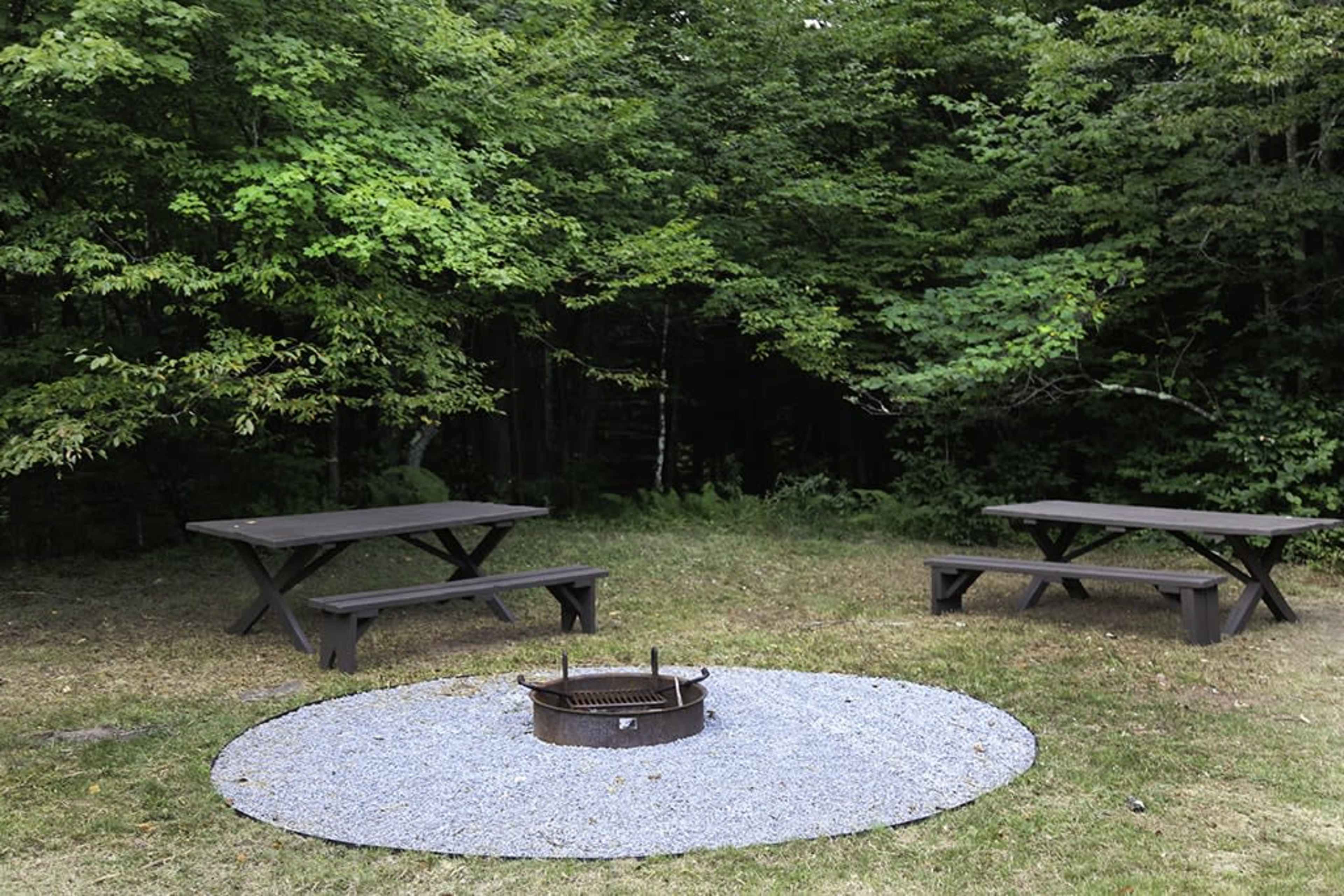 A clearing in a forest features two wooden picnic tables and a fire pit surrounded by a ring of gravel.