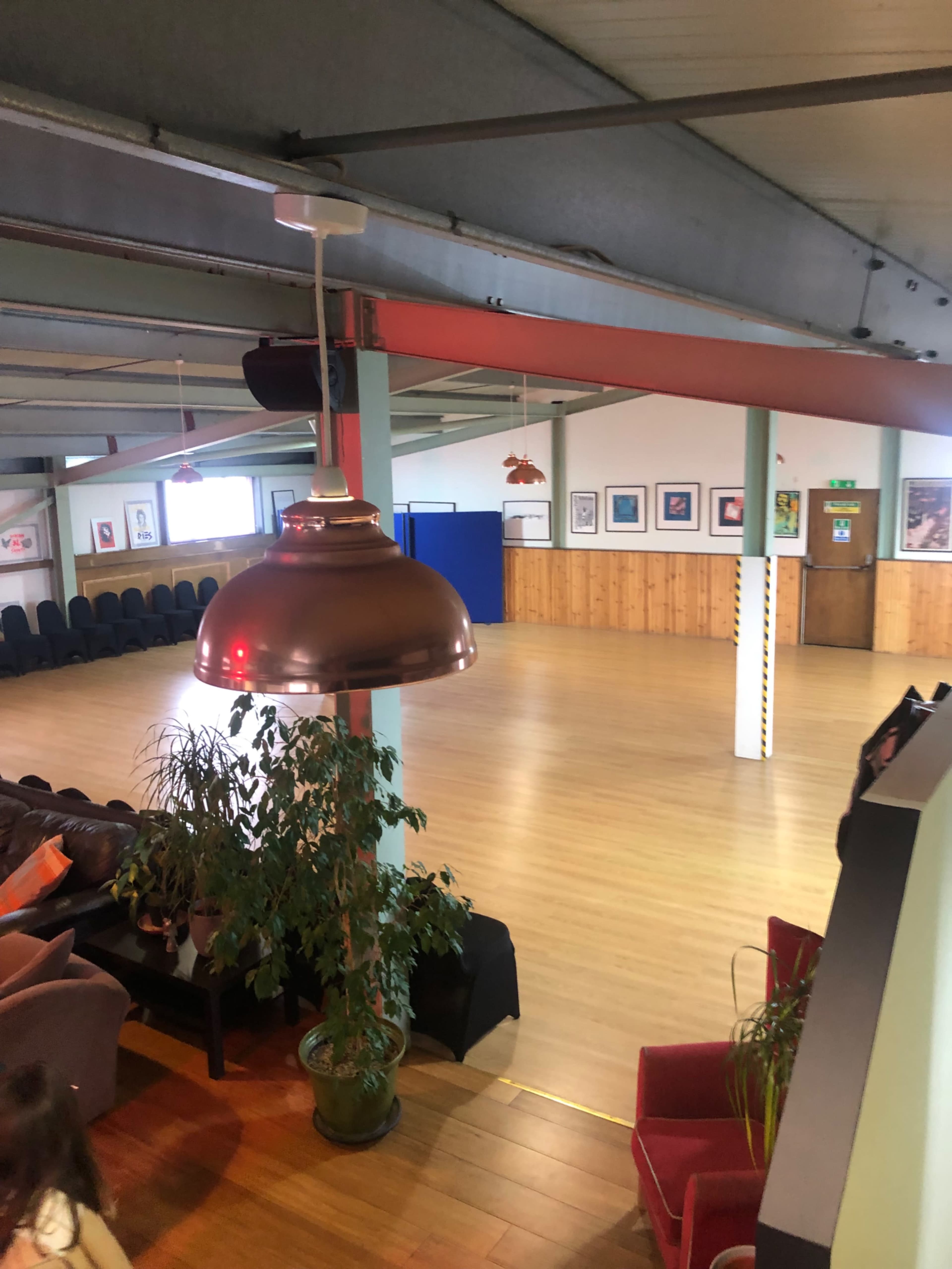 A spacious indoor hall with wooden flooring, seating arrangements along the walls, and decorative plants.