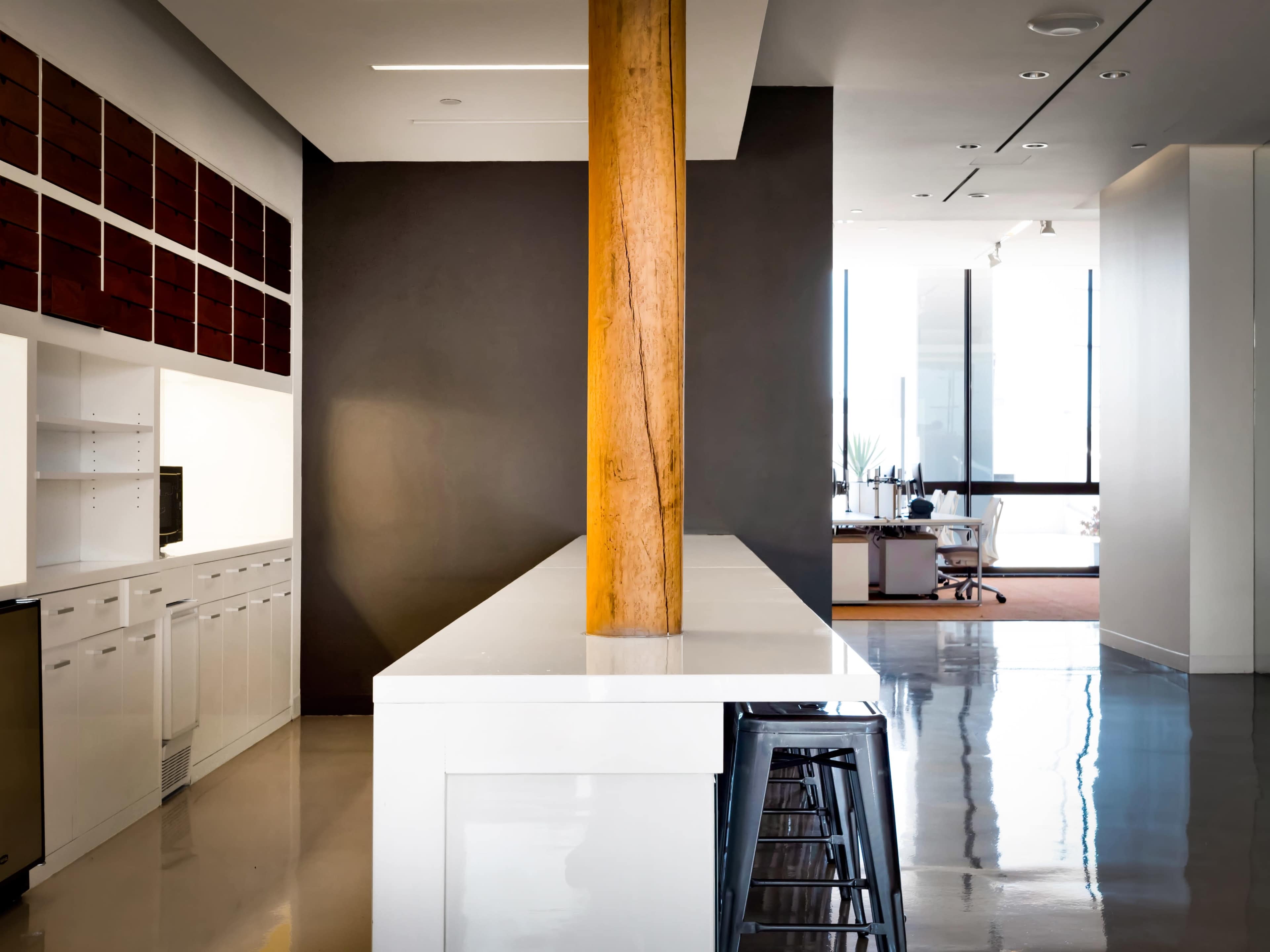 A modern kitchen area with a white countertop, a wooden column, and a workspace in the background featuring a desk and large windows.