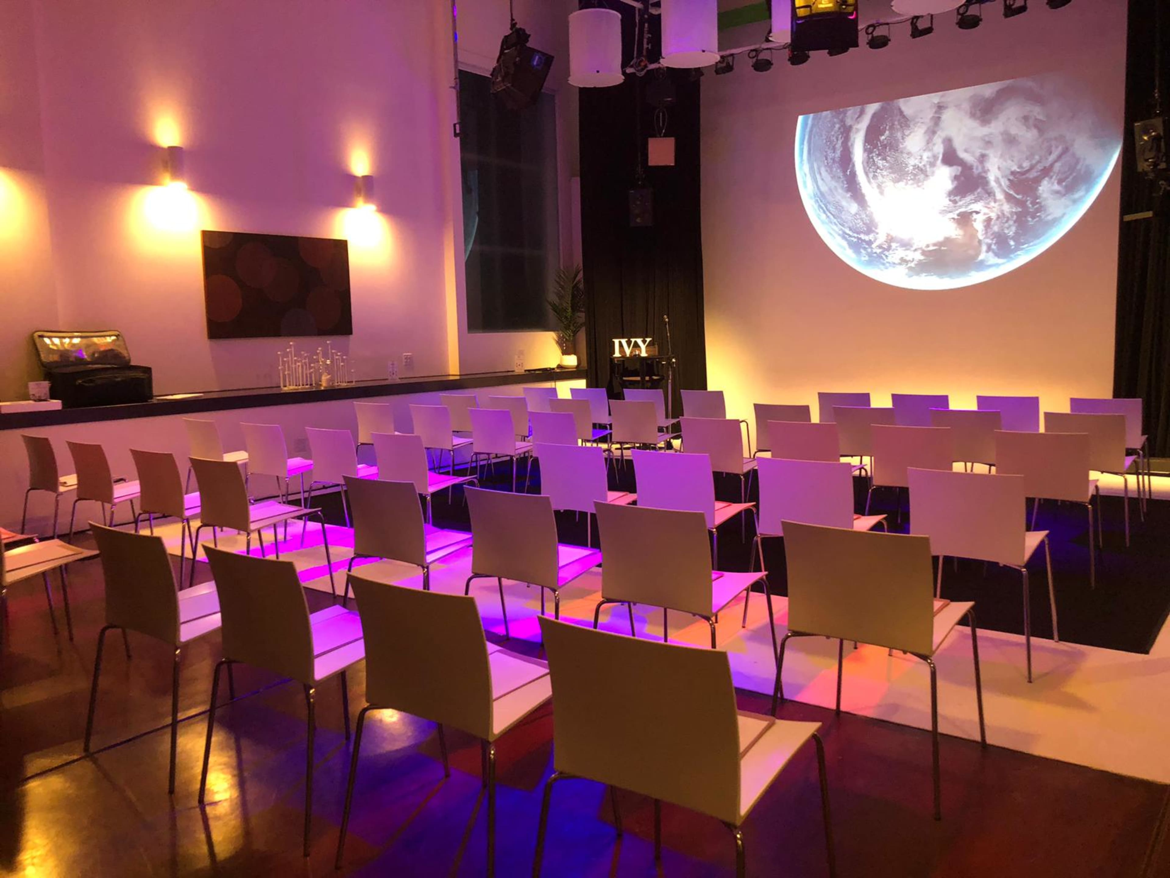 A room is set up for a presentation, featuring rows of white chairs facing a large projection of the Earth on a screen, with colored lighting and a small table in the corner.