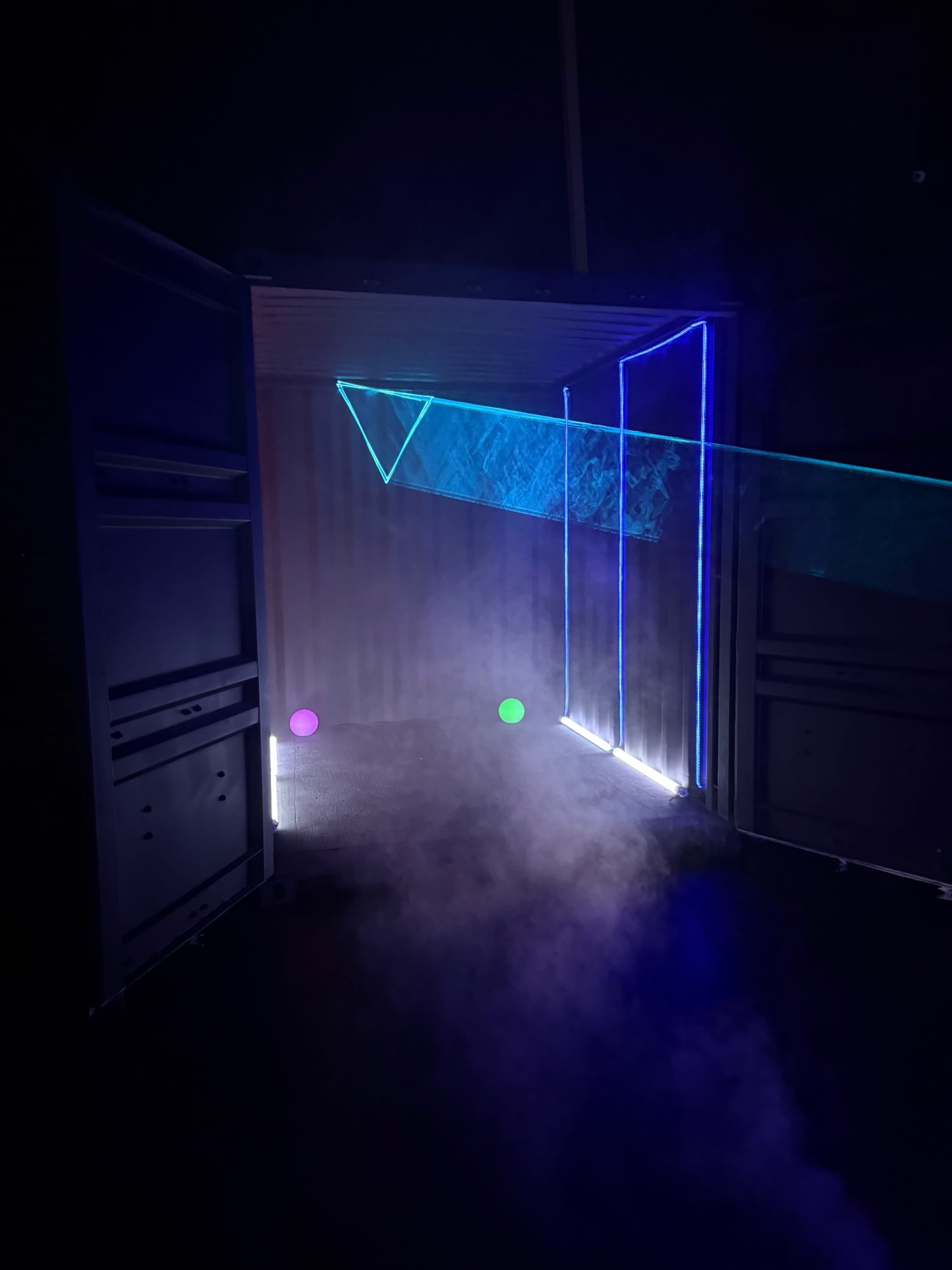 A dimly lit space features an open container illuminated by colorful lasers and smoke, creating geometric patterns on the walls and floor.