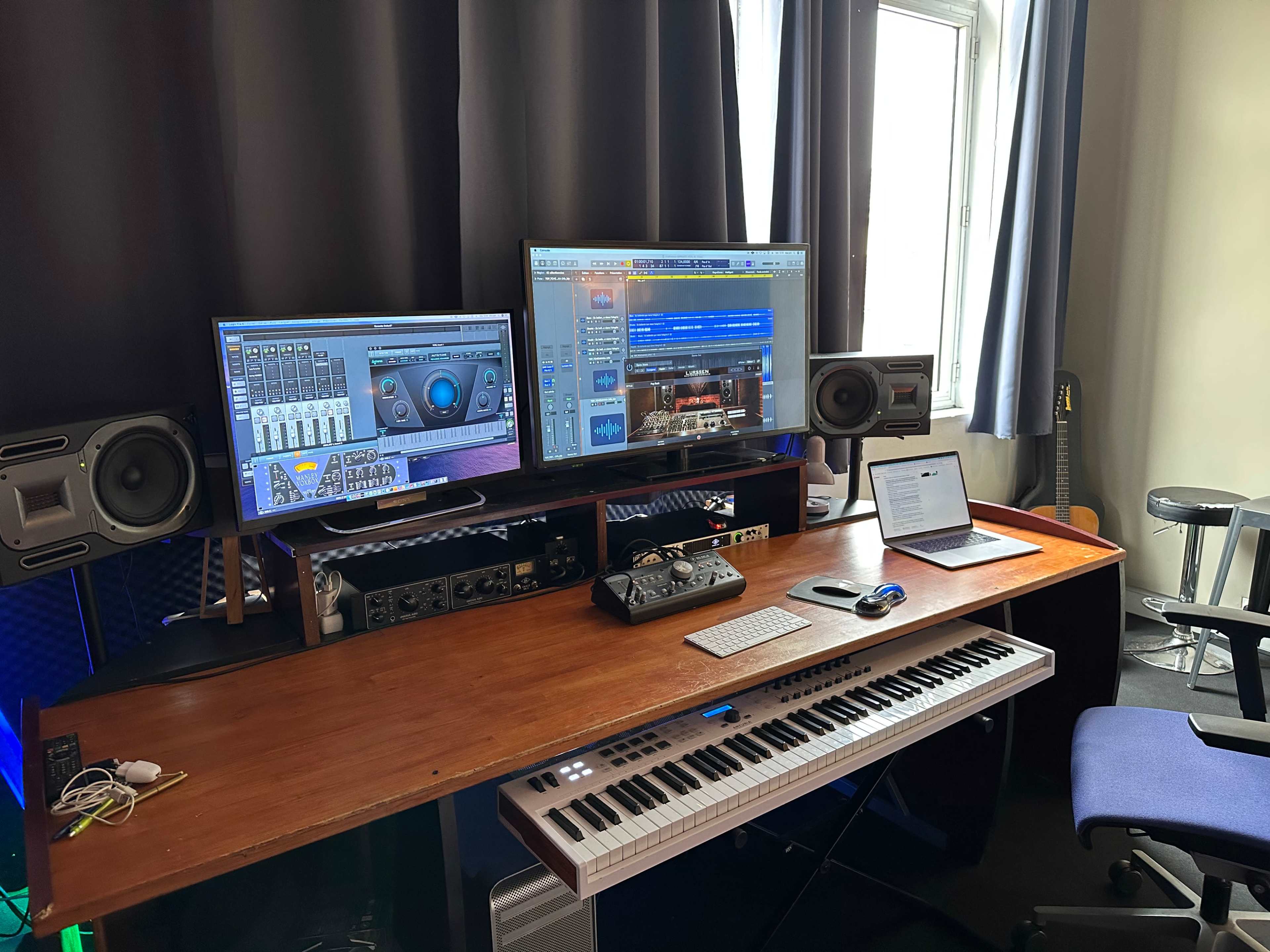 A music production studio is equipped with dual monitors, speakers, a keyboard, and a laptop on a wooden desk.