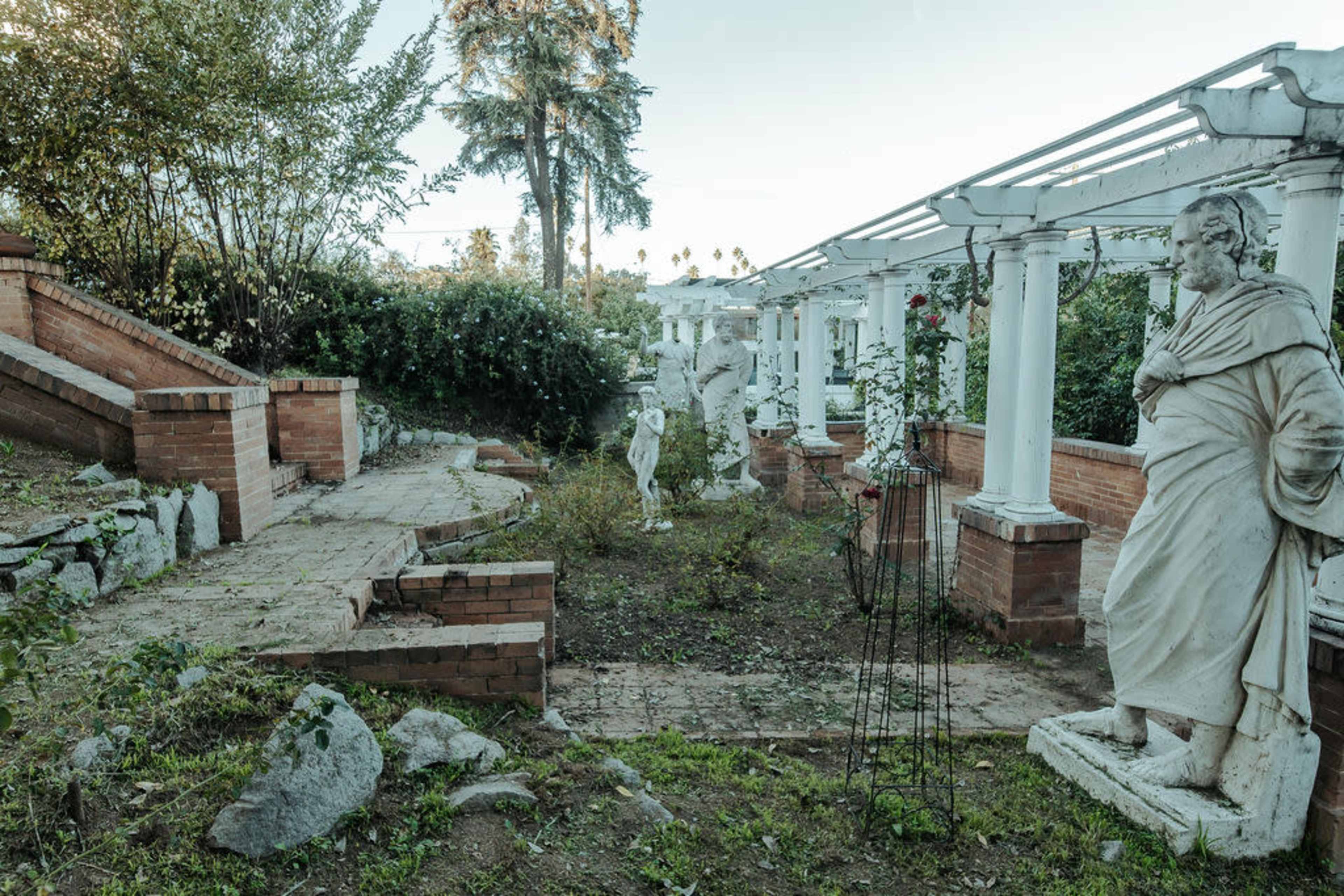 The image shows a landscaped area featuring a pathway flanked by stone statues and columns, with overgrown greenery surrounding the site.
