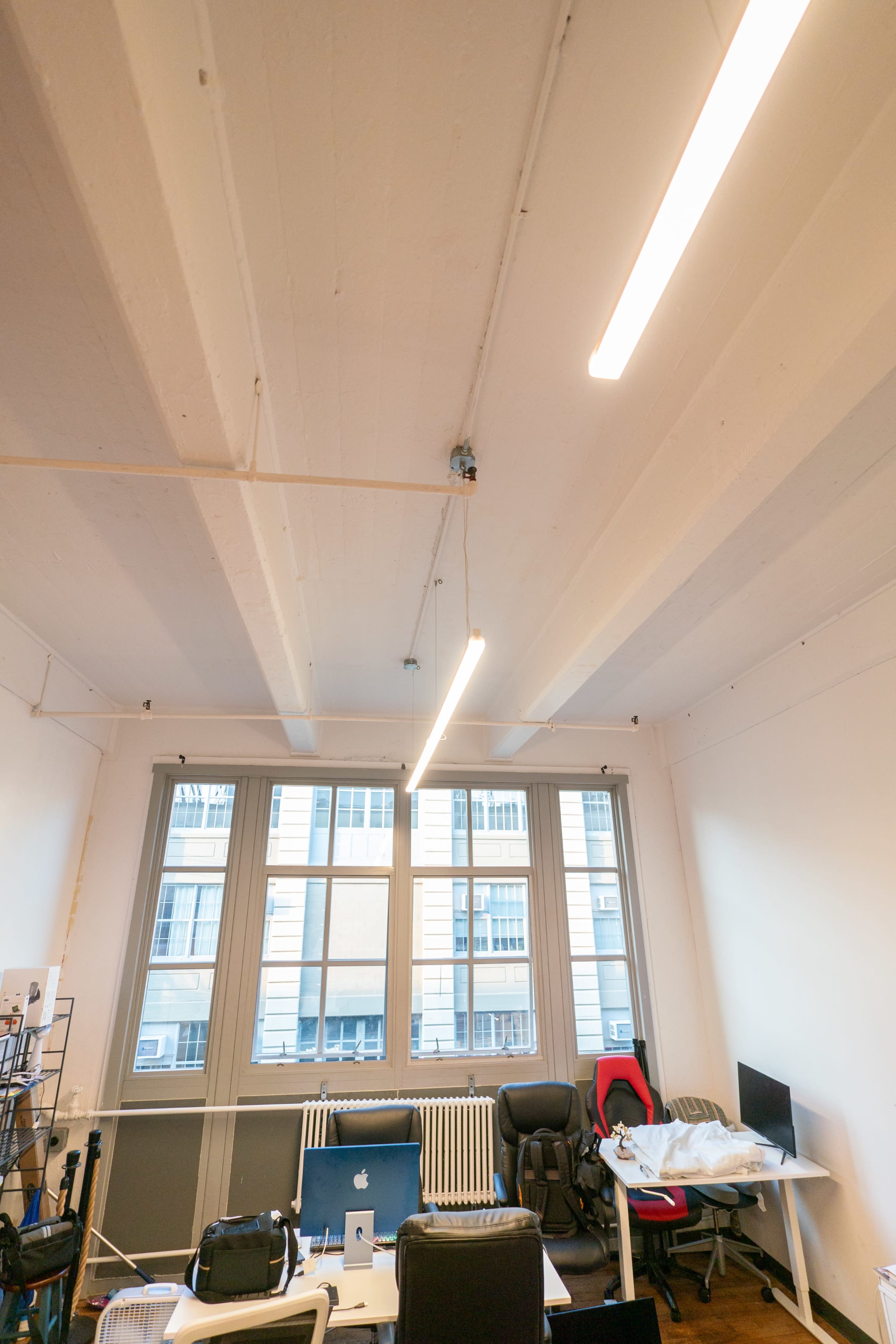 An interior office space with overhead lights, large windows, and various office furniture, including desks and chairs.
